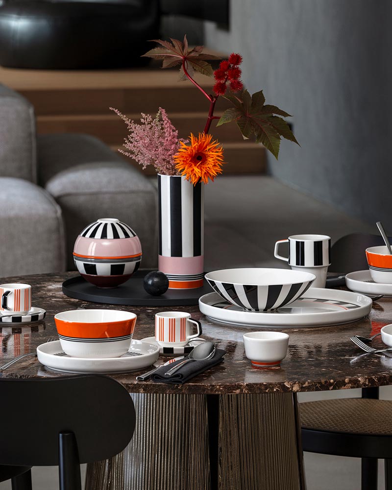 A dining table set with striped and colorful dishes and cups, featuring a decorative vase with vibrant flowers.