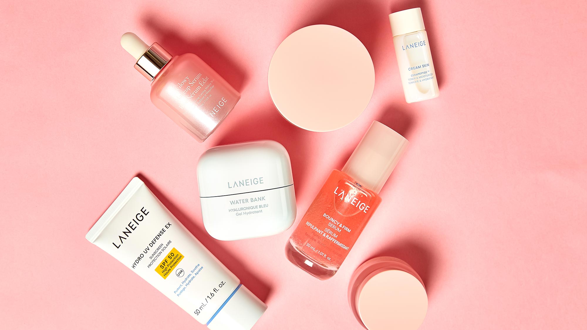 Skincare products arranged on a pink background, including creams, a serum, and a tube.