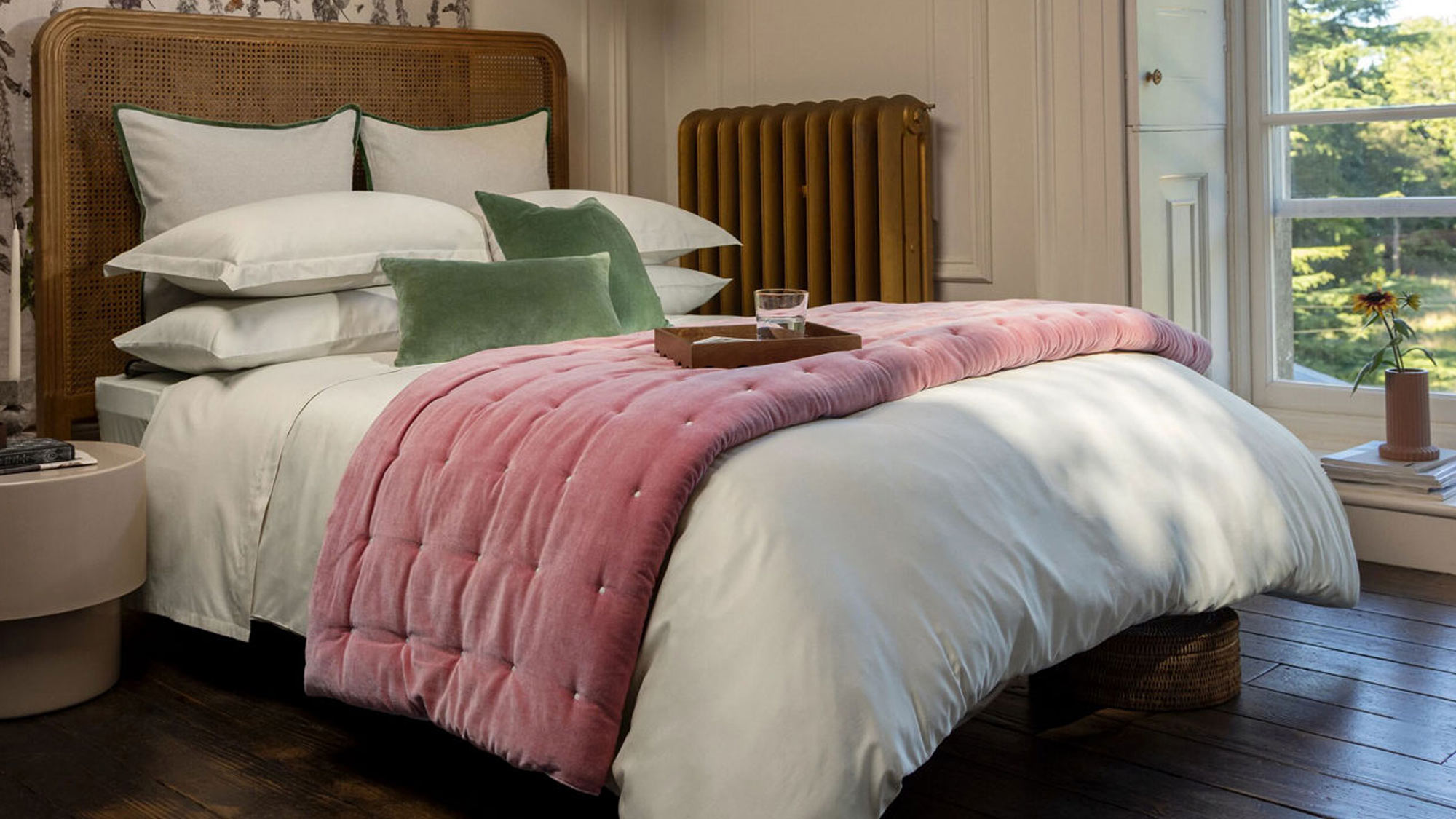 Cozy bedroom with a cane headboard, layered with white and green pillows, a pink quilt, wooden floor, and a large window with a garden view.