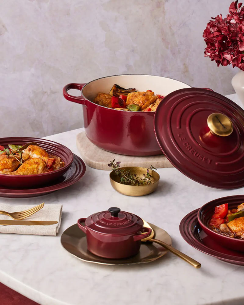 Red cookware with food, arranged on a marble table with plates and silverware. A pot of flowers decorates the background.