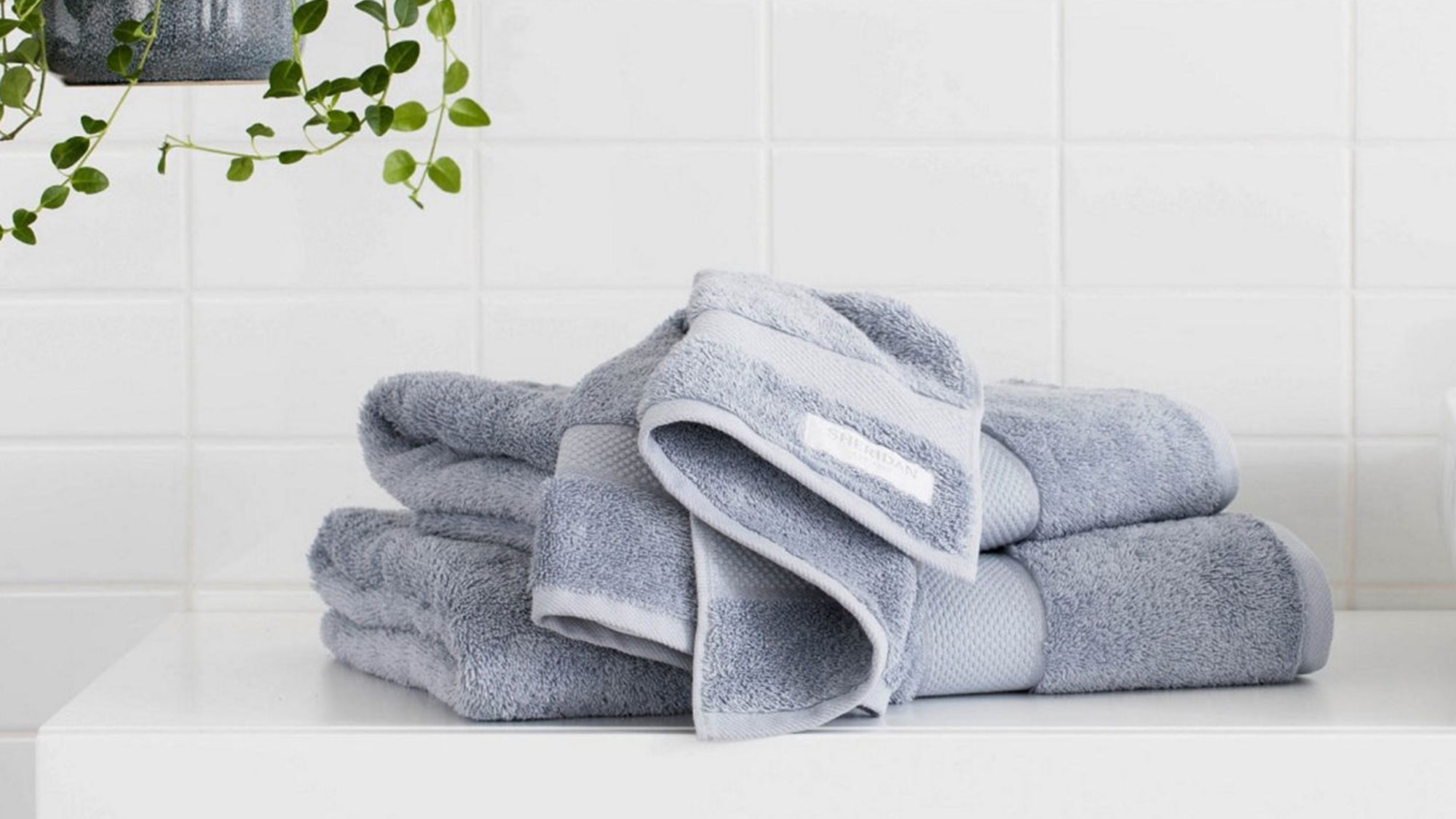 Gray towels stacked on a white countertop with a hanging plant and white tiled wall.