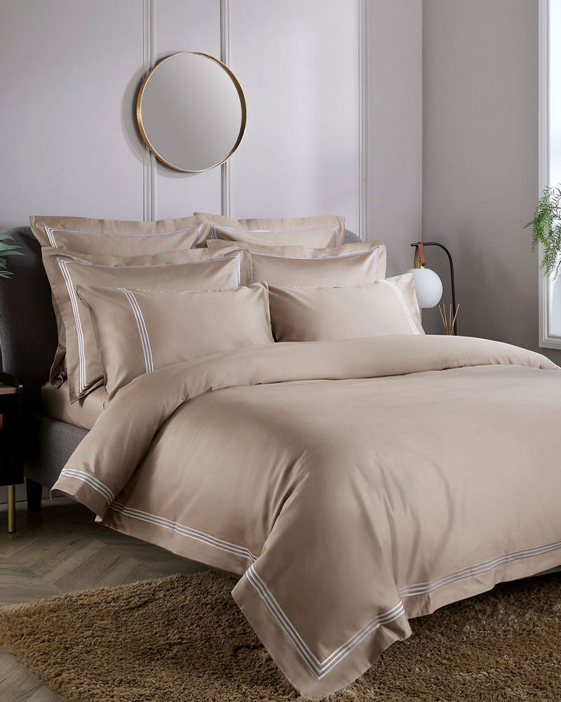 A neatly made bed with beige linens, flanked by plants and a round mirror on the wall, in a softly lit bedroom.