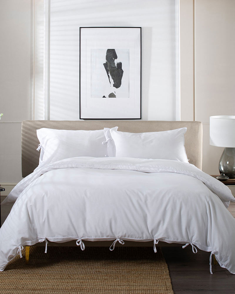 A neatly made bed with white linens is centered in a minimalistic bedroom. There's abstract art above the headboard and side tables with lamps.