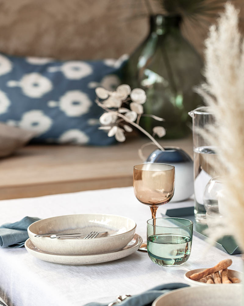 Elegant table setting with neutral dishware, blue napkins, a glass tumbler, and dried flowers. Background has patterned cushions and a vase.