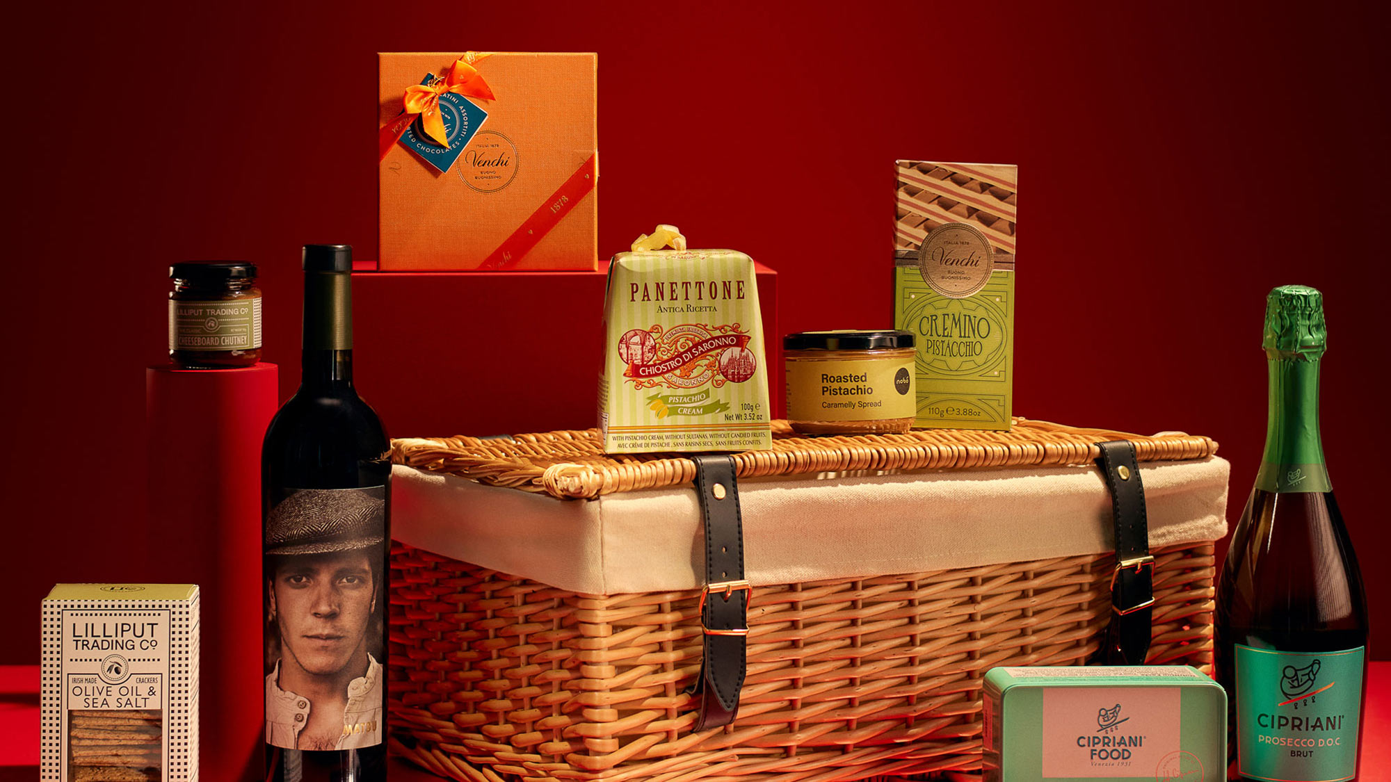 A wicker basket surrounded by gourmet food and wine items, set against a red background.