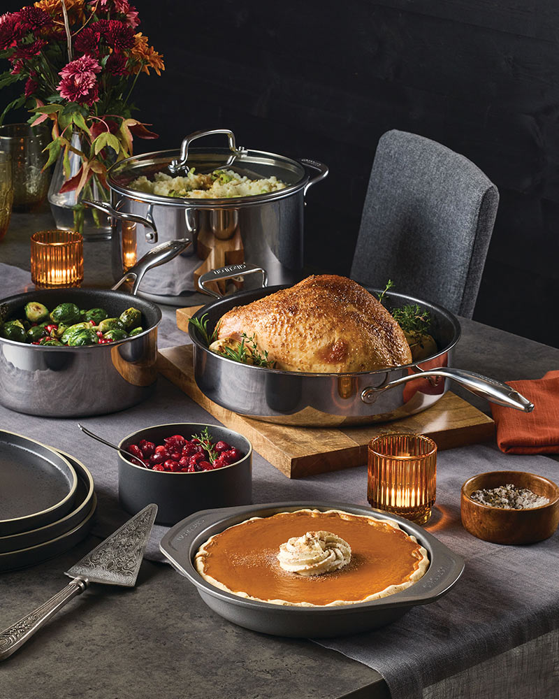 A table set with Thanksgiving dishes, including a roast turkey, Brussels sprouts, cranberries, a pie, flowers, and candles.