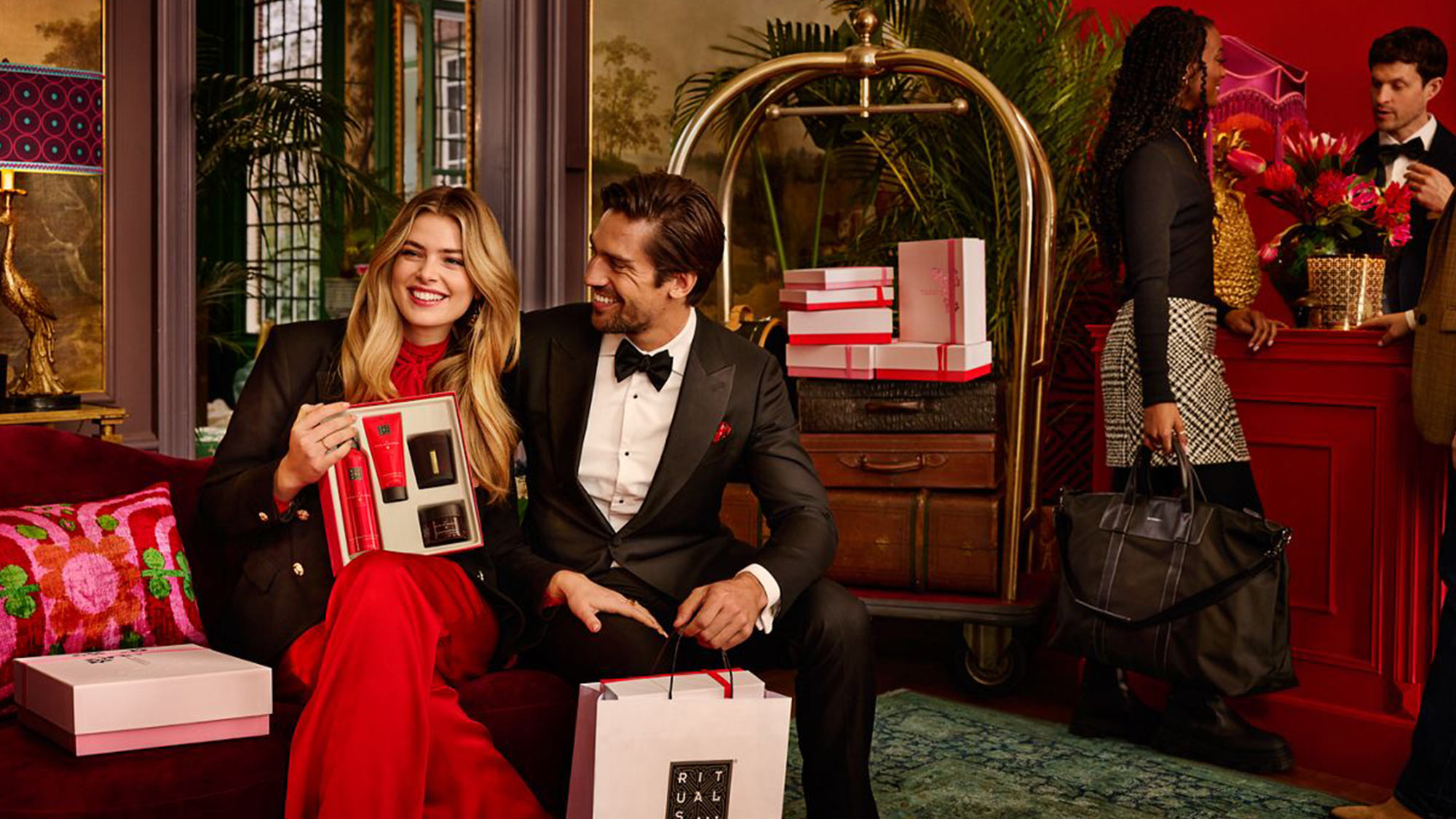 A happy couple, dressed elegantly, sit with gift boxes in a festive setting. A bellhop and a woman in the background add to a luxurious atmosphere.