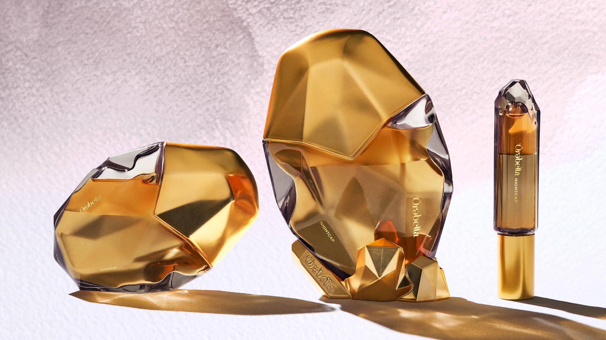 Three gold-toned, gem-shaped perfume bottles on a textured surface with soft pink lighting.