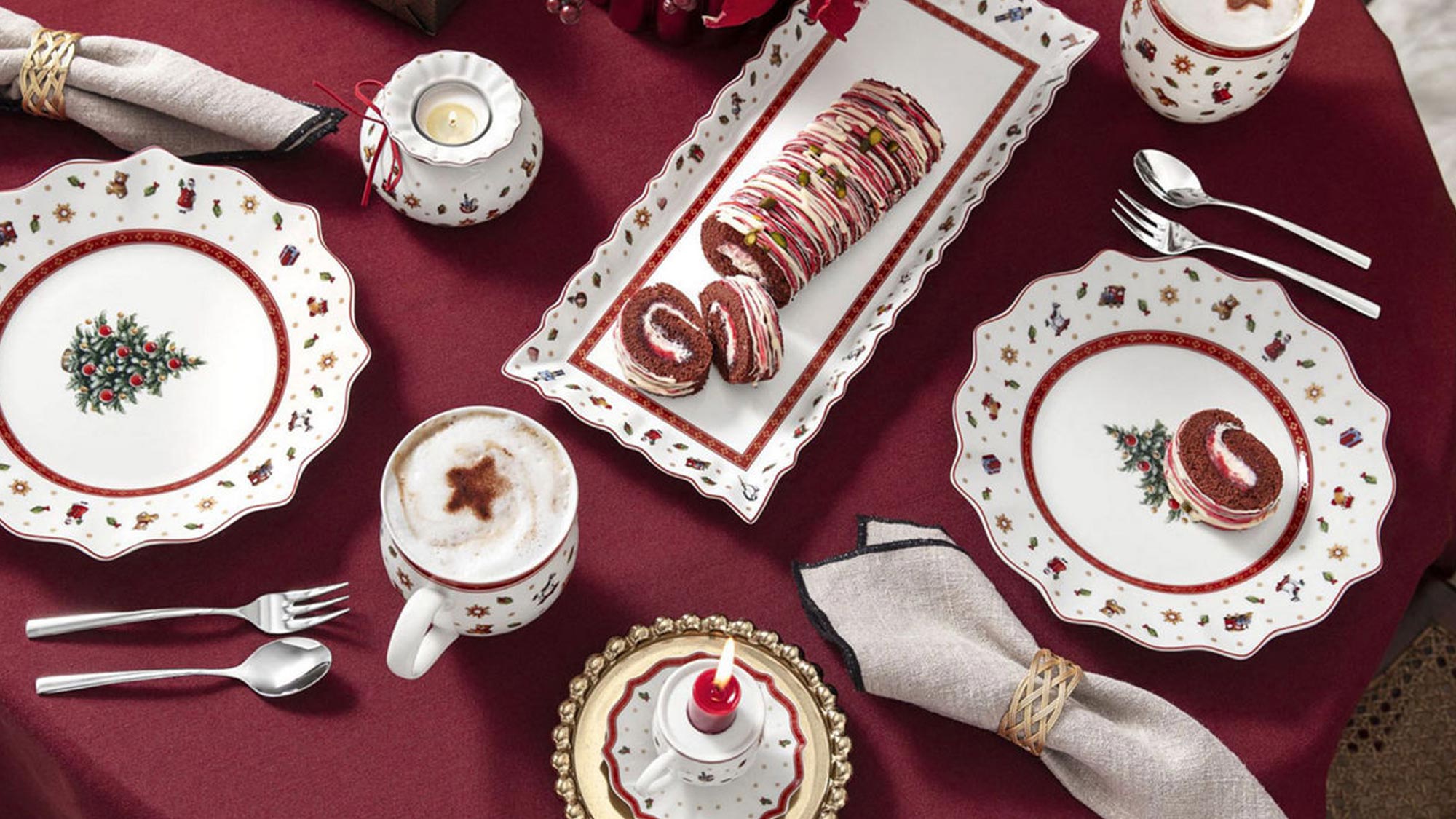 Elegant holiday table with red cloth, festive plates, mugs, cutlery, napkins, pastries, a gift, and poinsettias centerpiece.