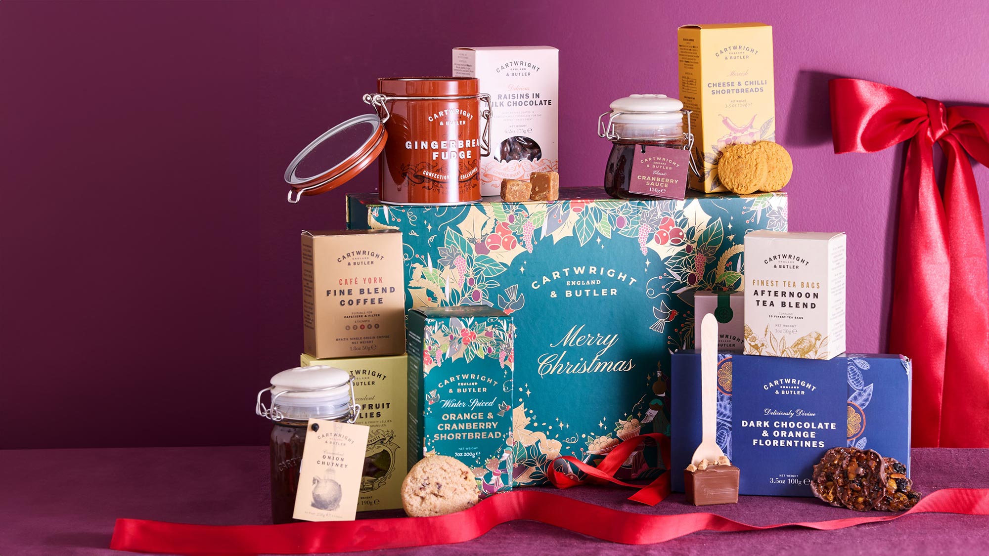 A festive gift hamper with tea, biscuits, chocolates, and jars, set against a purple background with red ribbon decorations.