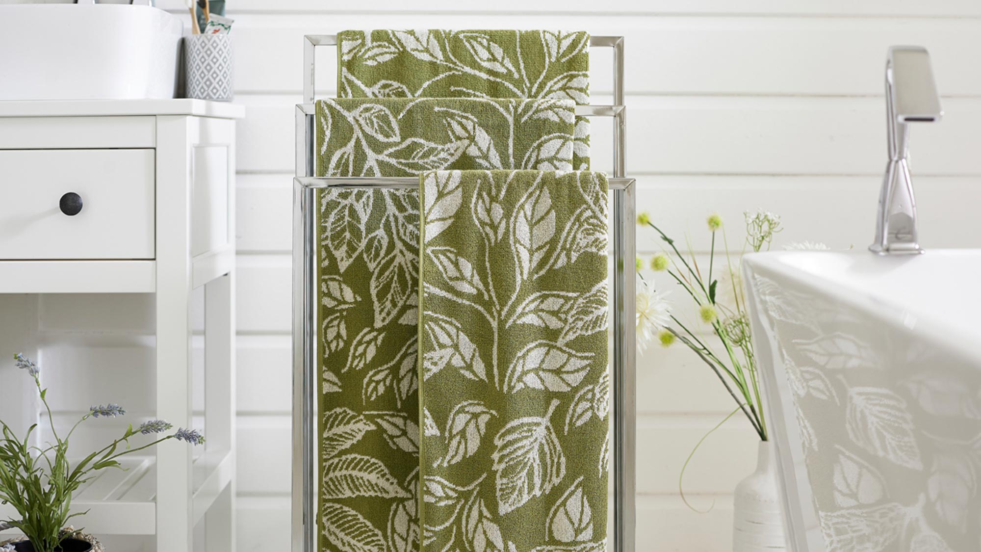 Bathroom with a towel rack holding green leaf-patterned towels, a white sink cabinet, and decorative plants on a white tiled floor.