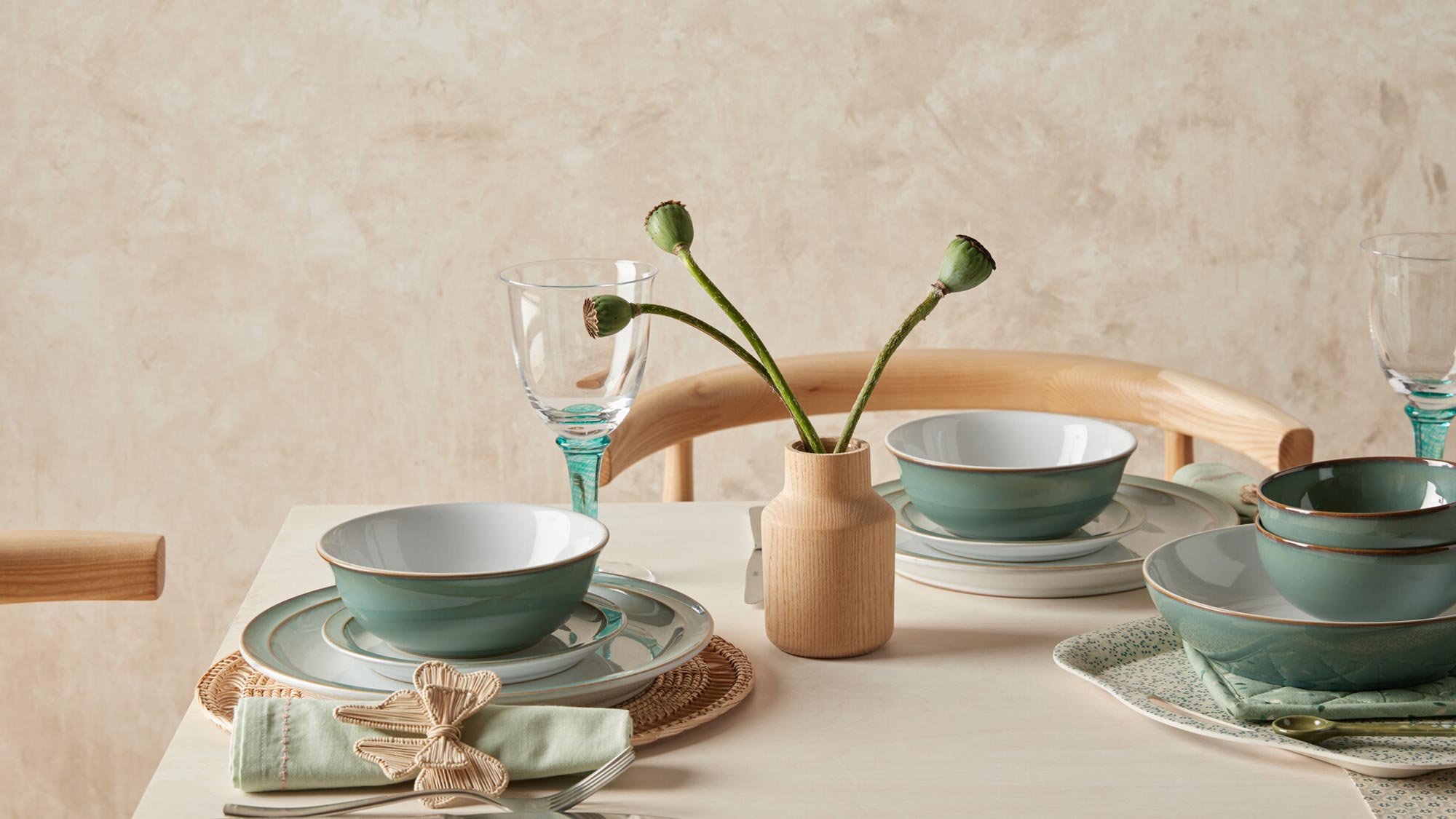 A minimalist table setting with green dishes, glasses, a small vase with buds, and neatly arranged napkins on a light wood table.