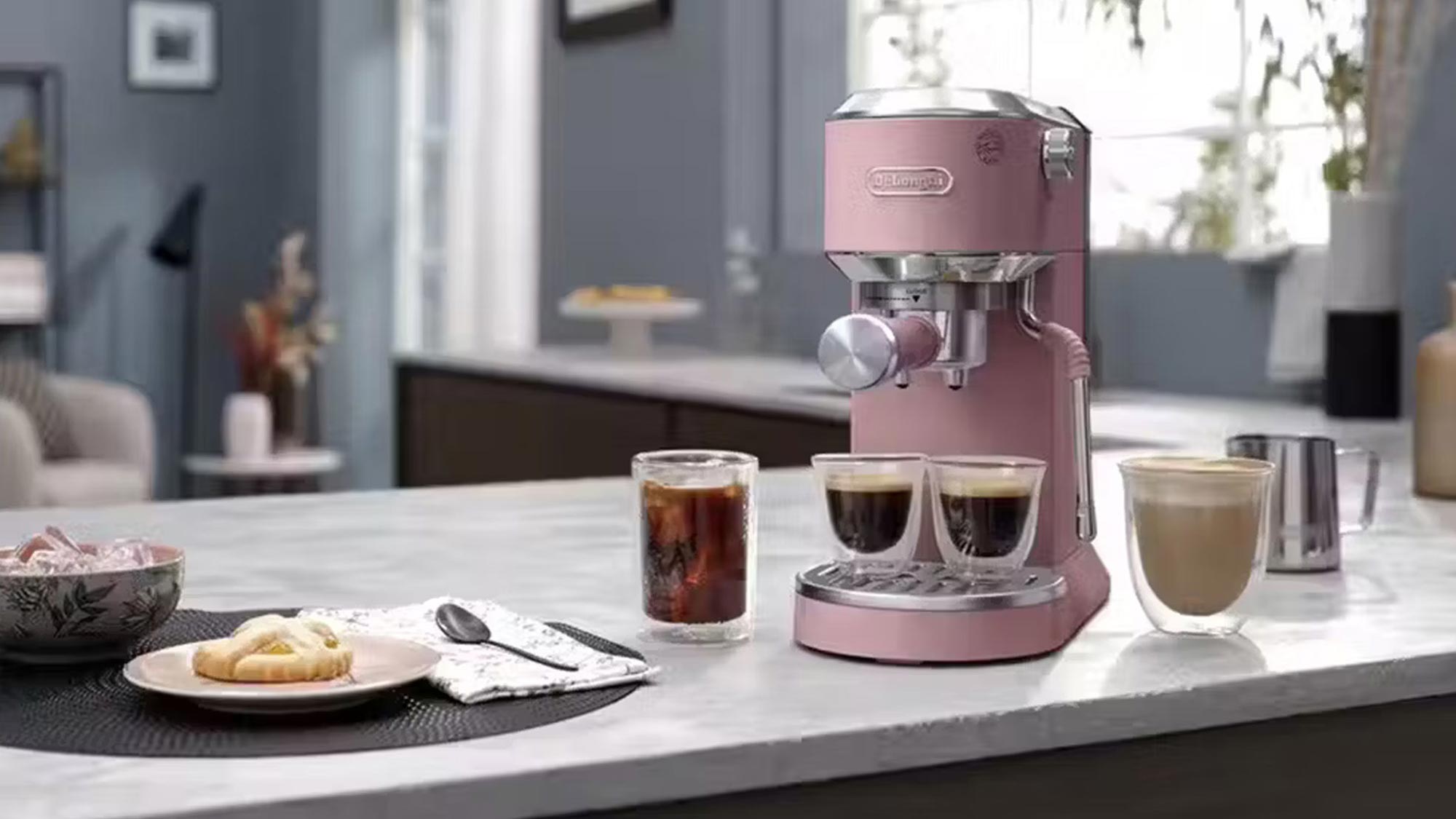 A pink espresso machine on a kitchen counter with coffee and pastries nearby.