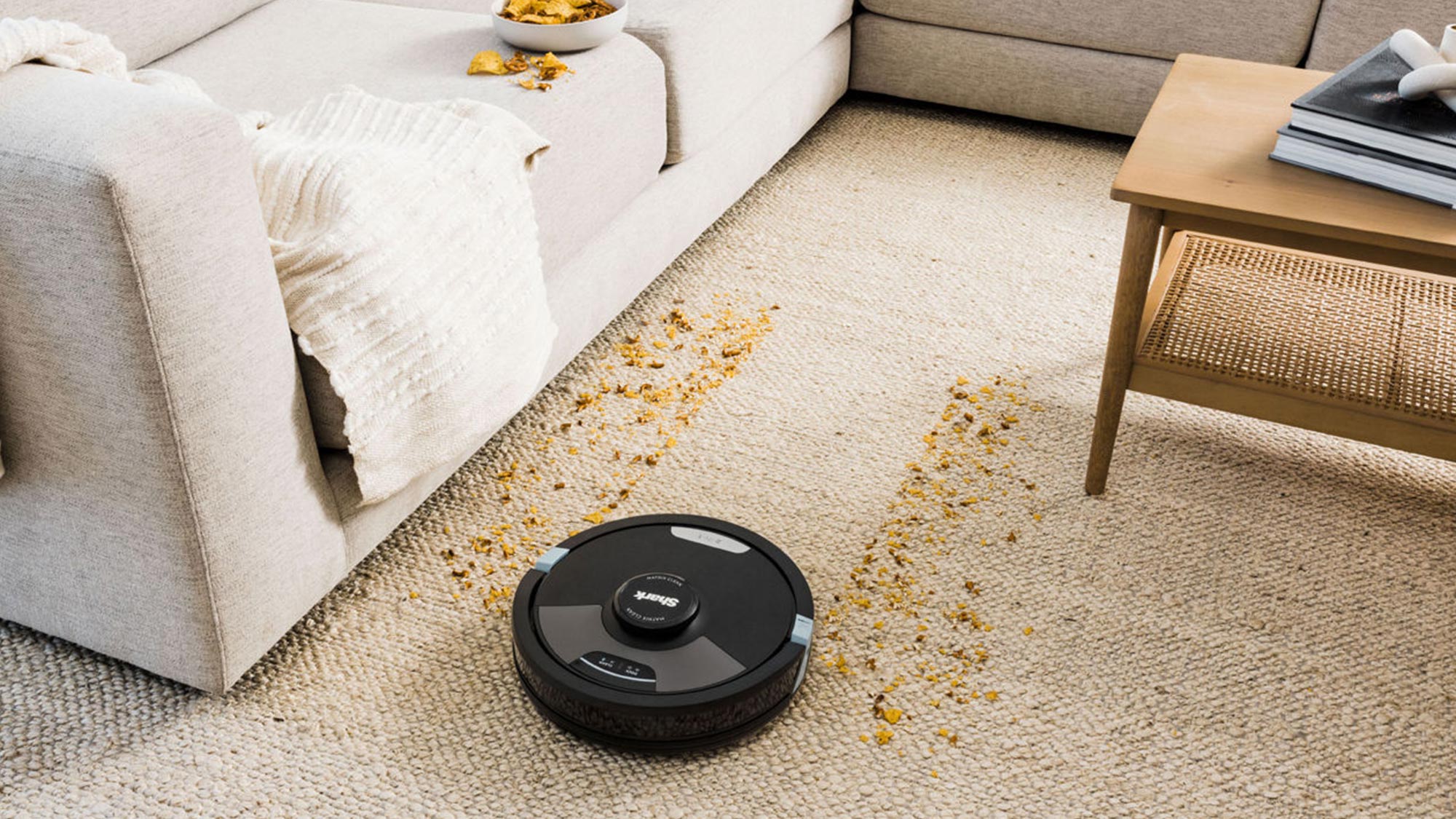 A robotic vacuum cleaner is cleaning crumbs on a carpet in a living room with a sofa and a coffee table.