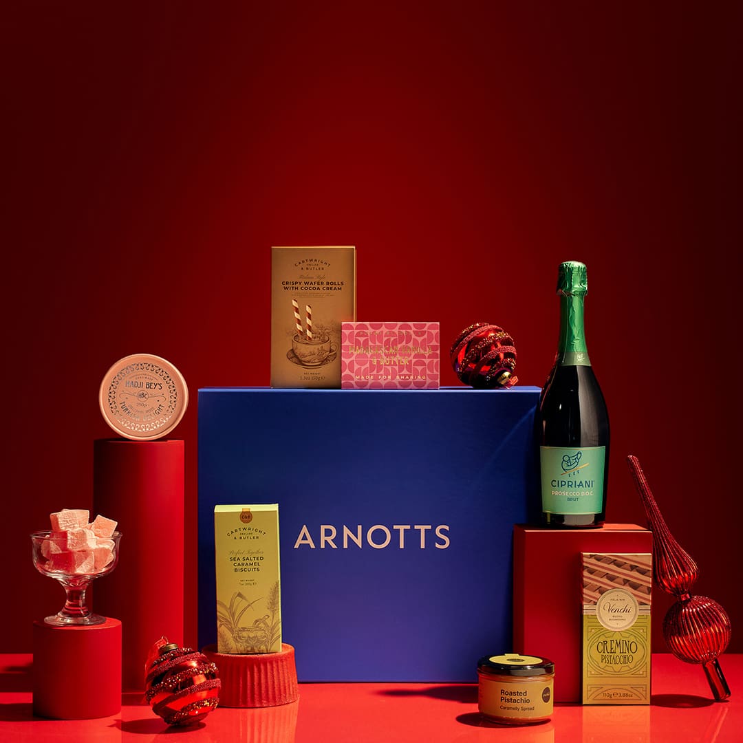 A collection of luxury food items, including champagne and sweets, arranged around a blue Arnott's gift box on a red backdrop.