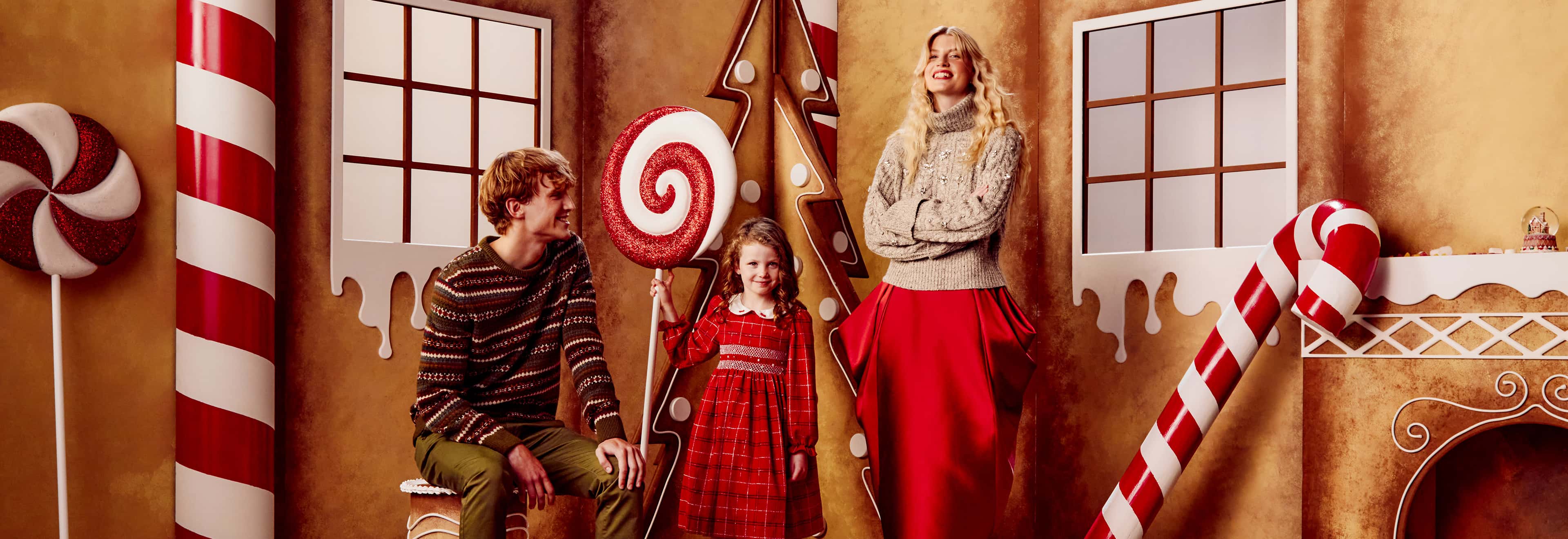 Three people in festive attire stand in a gingerbread house setting with large candy decorations.