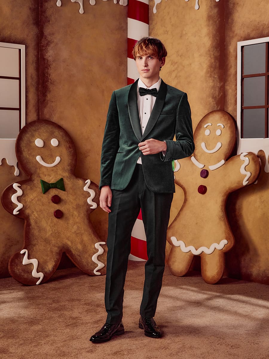 A person in a green velvet suit stands between two large gingerbread figures, set against a festive backdrop.