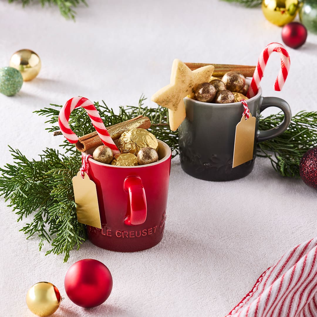 Two festive mugs filled with gold-wrapped chocolates, candy canes, a star cookie, and cinnamon sticks, surrounded by Christmas decorations.