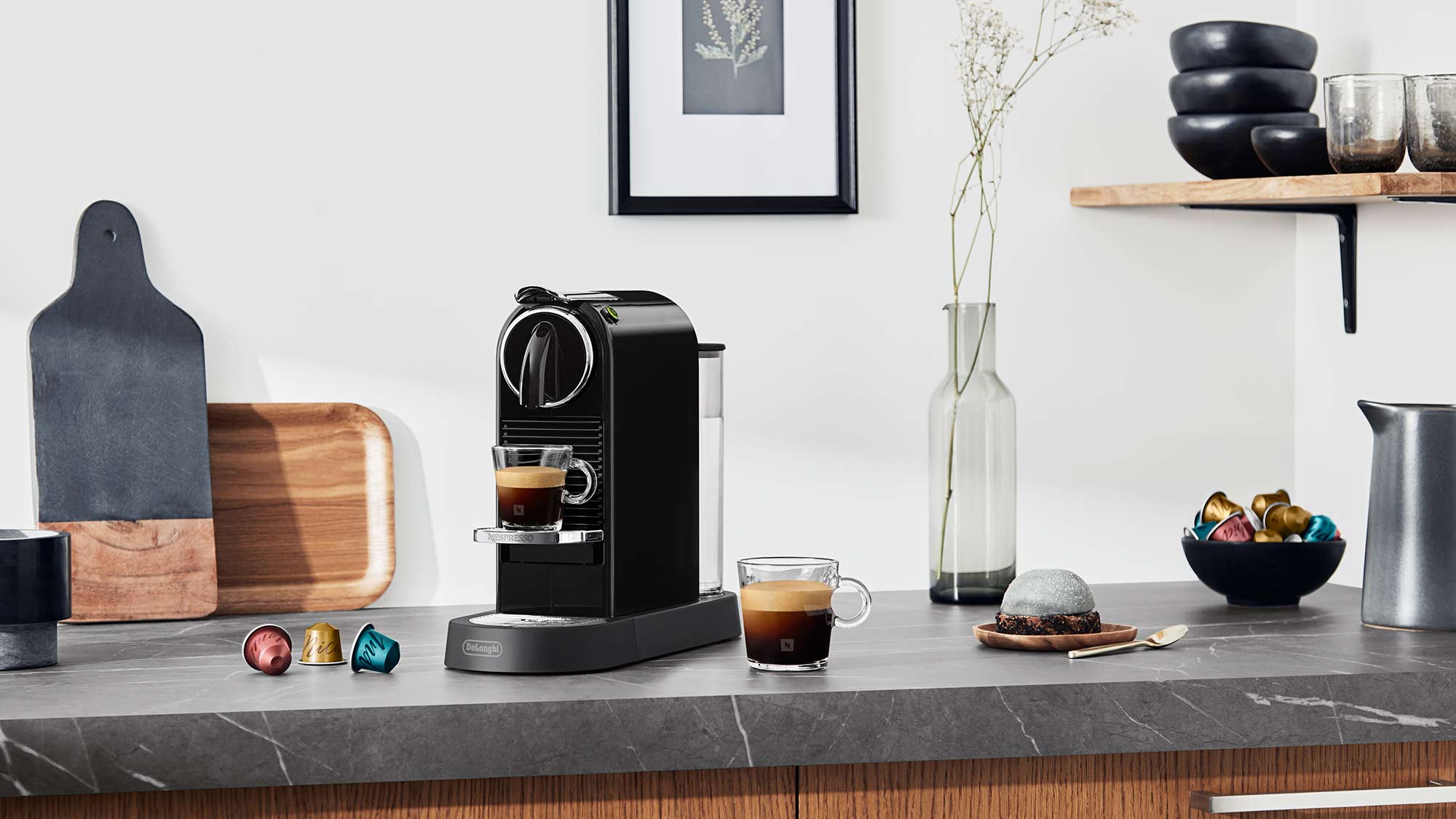 A modern kitchen countertop with a black espresso machine, coffee capsules, glassware, and decorative items on a wooden shelf above.