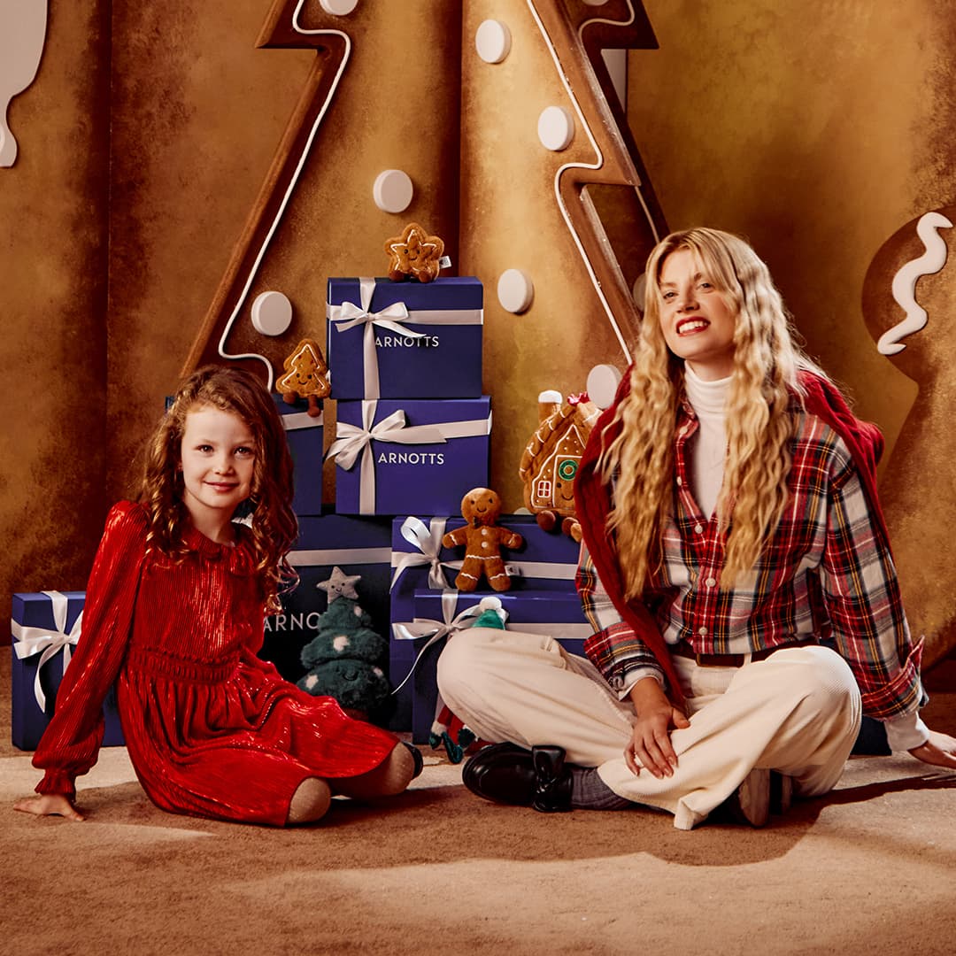 A woman and a young girl sit beside a stack of gift boxes in a festive room with a gingerbread-themed backdrop.