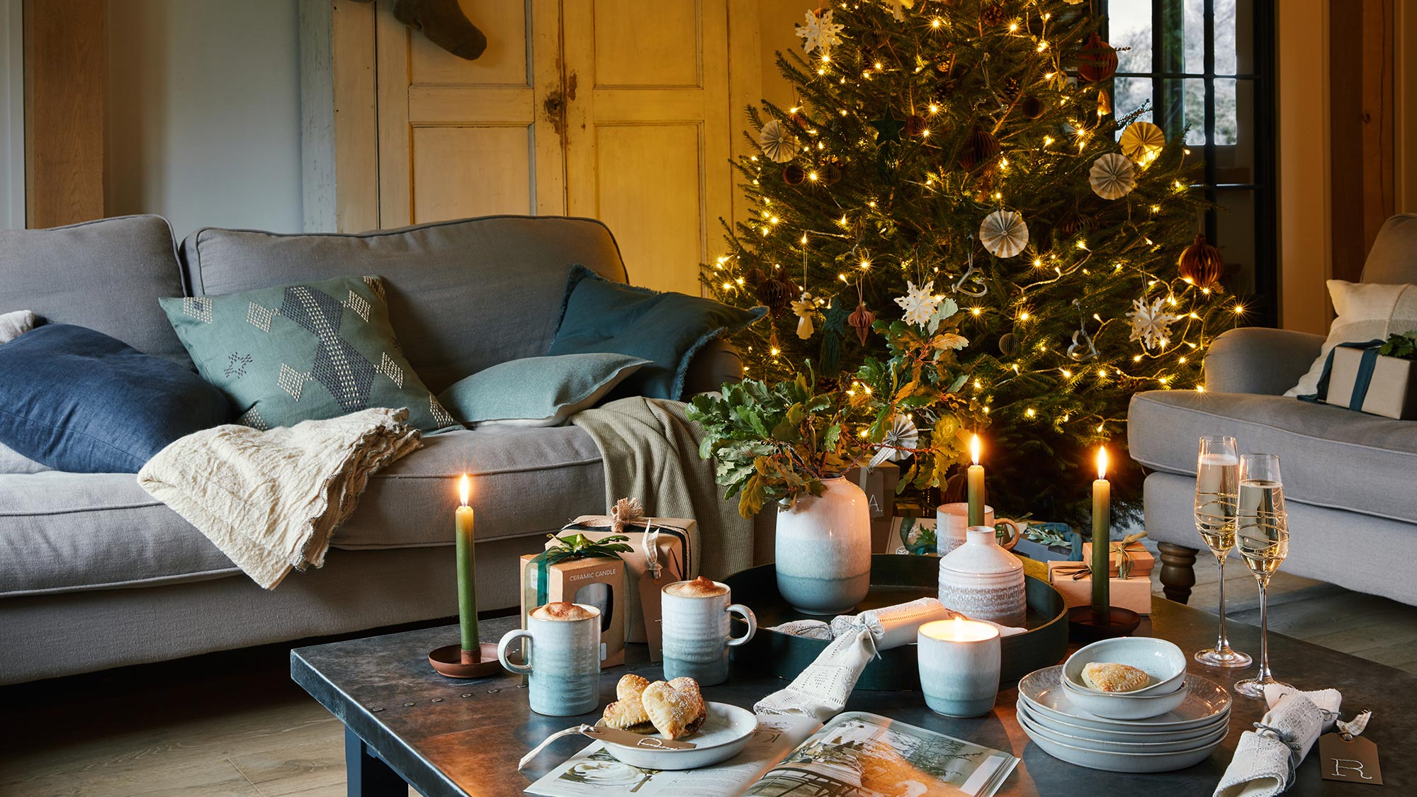 Cozy living room with a decorated Christmas tree, lit candles, and a coffee table set with cups and snacks.