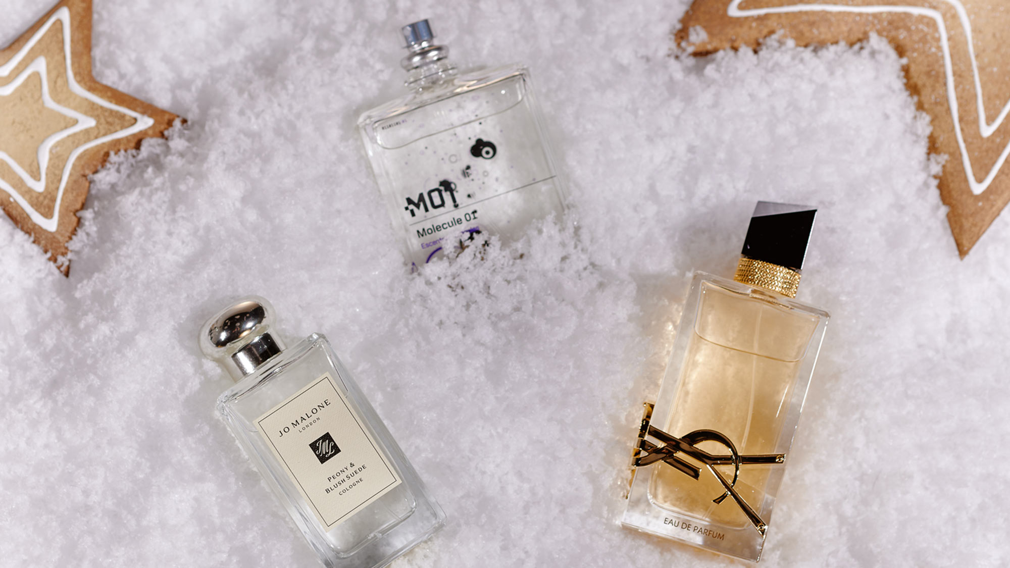 Three perfume bottles on snowy ground, surrounded by star-shaped gingerbread cookies.