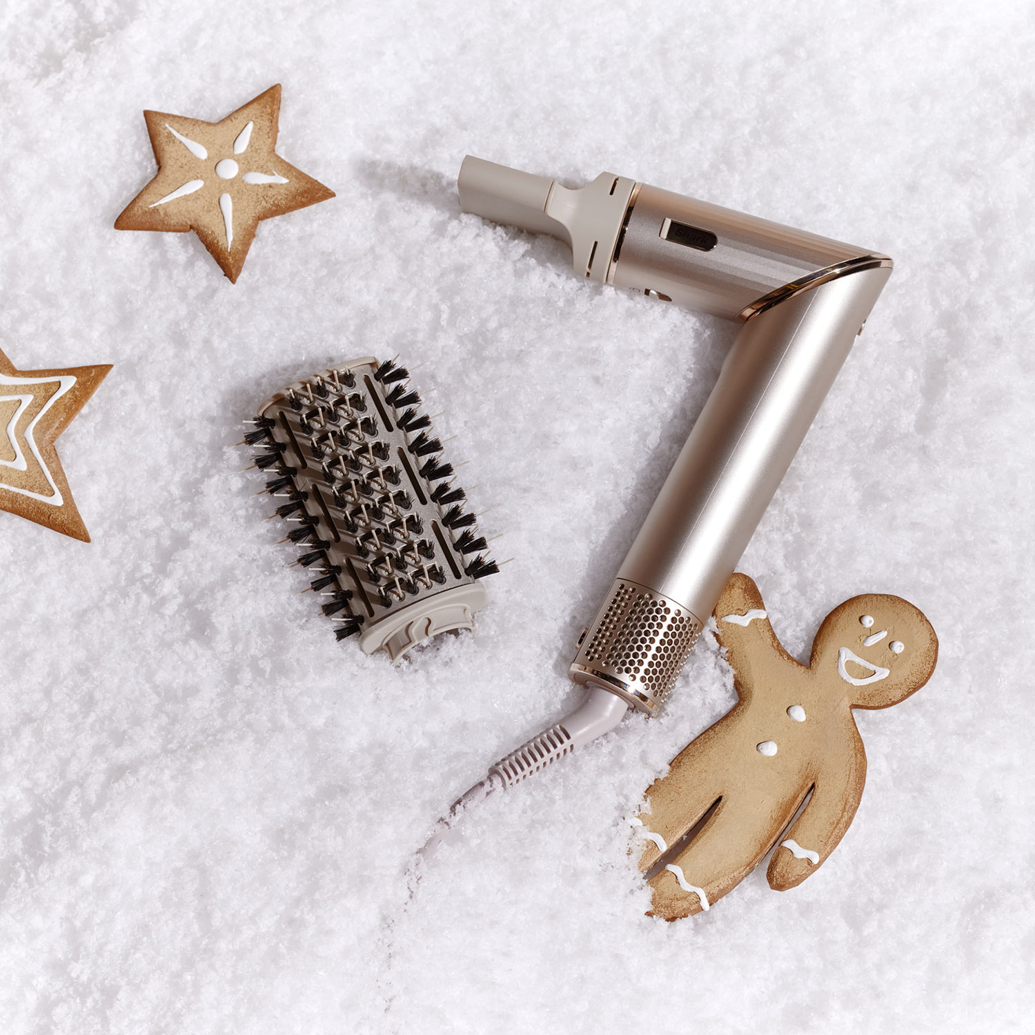 A metallic hair dryer and brush are on snowy white ground, surrounded by star-shaped and gingerbread cookies.