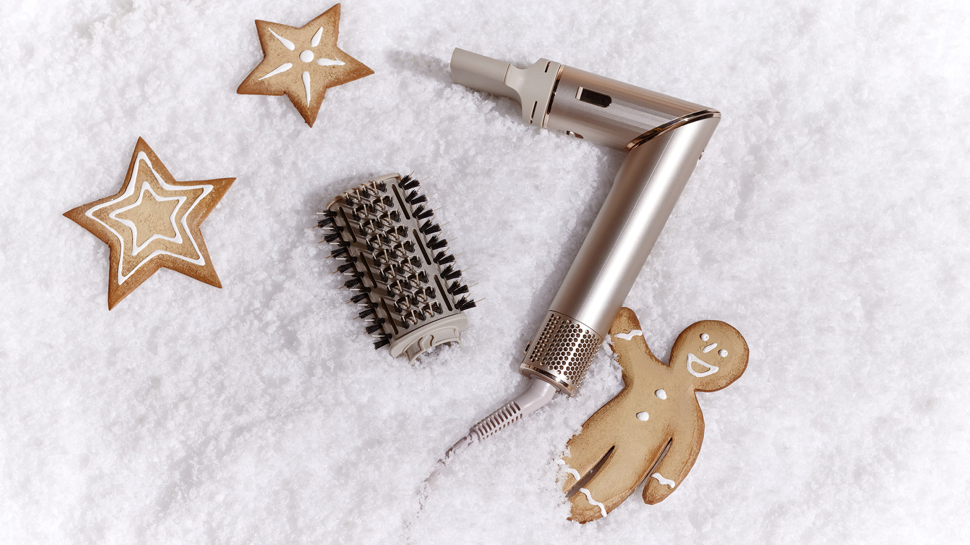 A metallic hair dryer and brush are on snowy white ground, surrounded by star-shaped and gingerbread cookies.