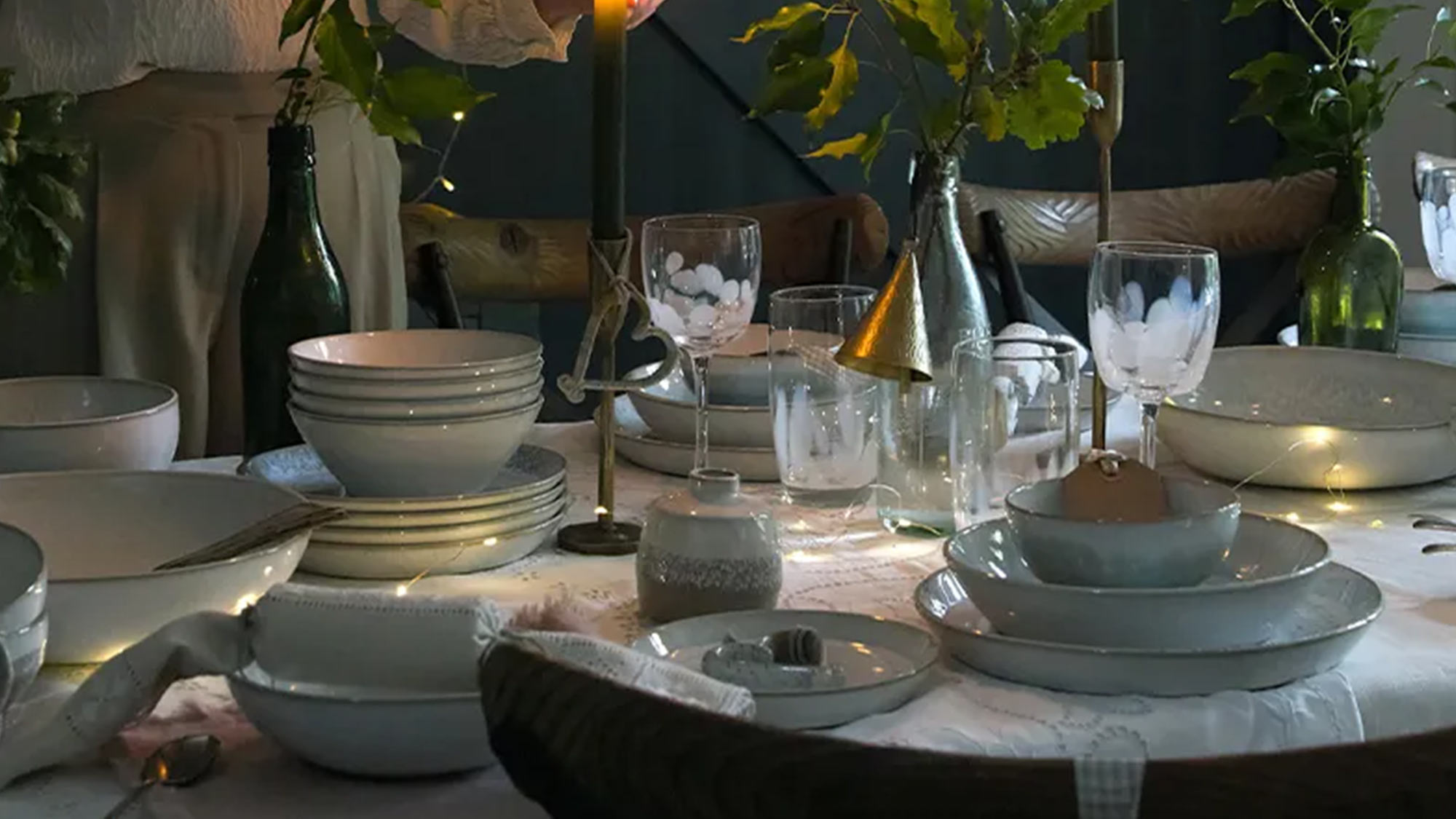 A dimly lit dining table is set with dishes, candles, and greenery. A person in a light blouse is adjusting a candle.