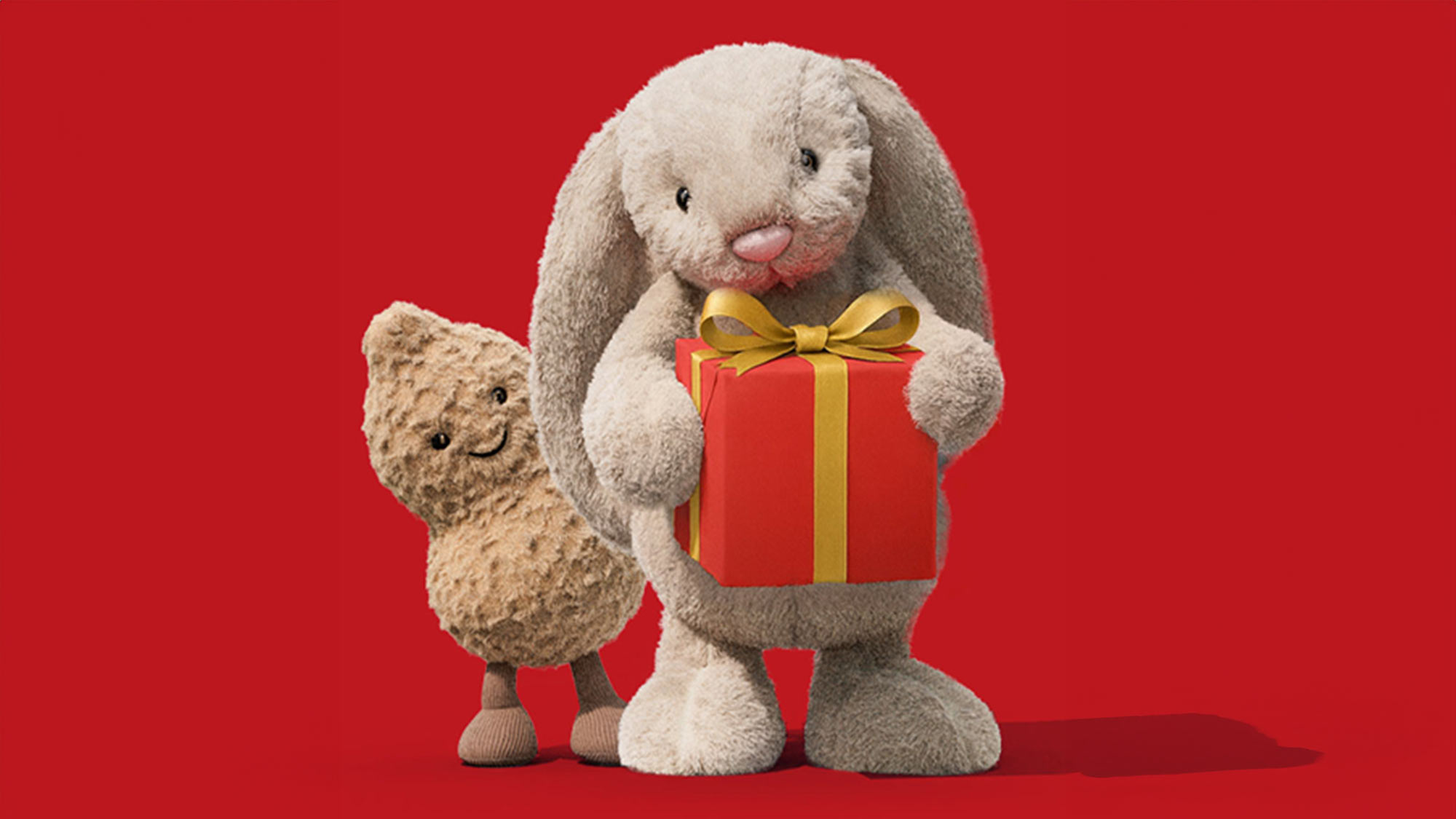 A plush bunny holding a red gift with a yellow ribbon stands next to a smiling plush peanut against a red background.
