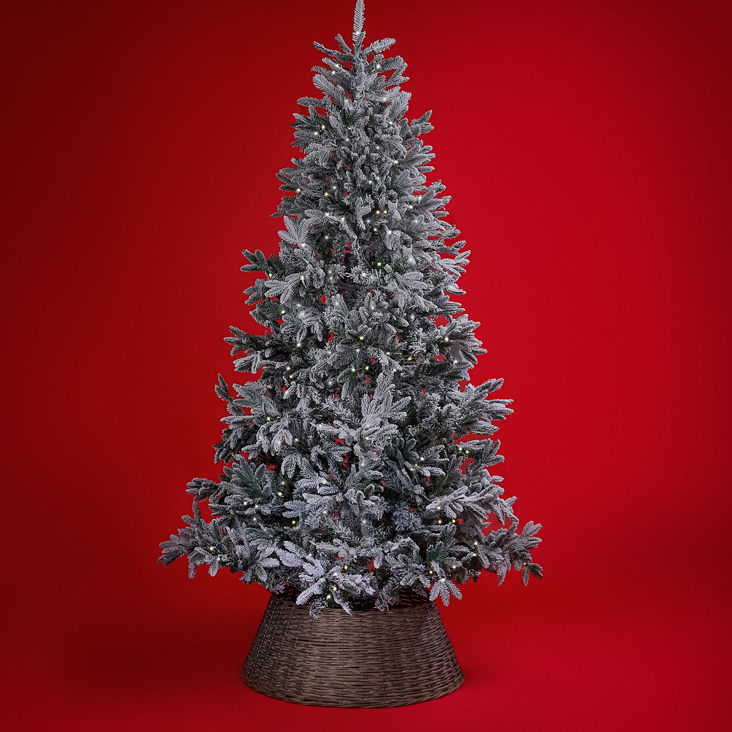 A snowy artificial Christmas tree with lights stands against a red background.