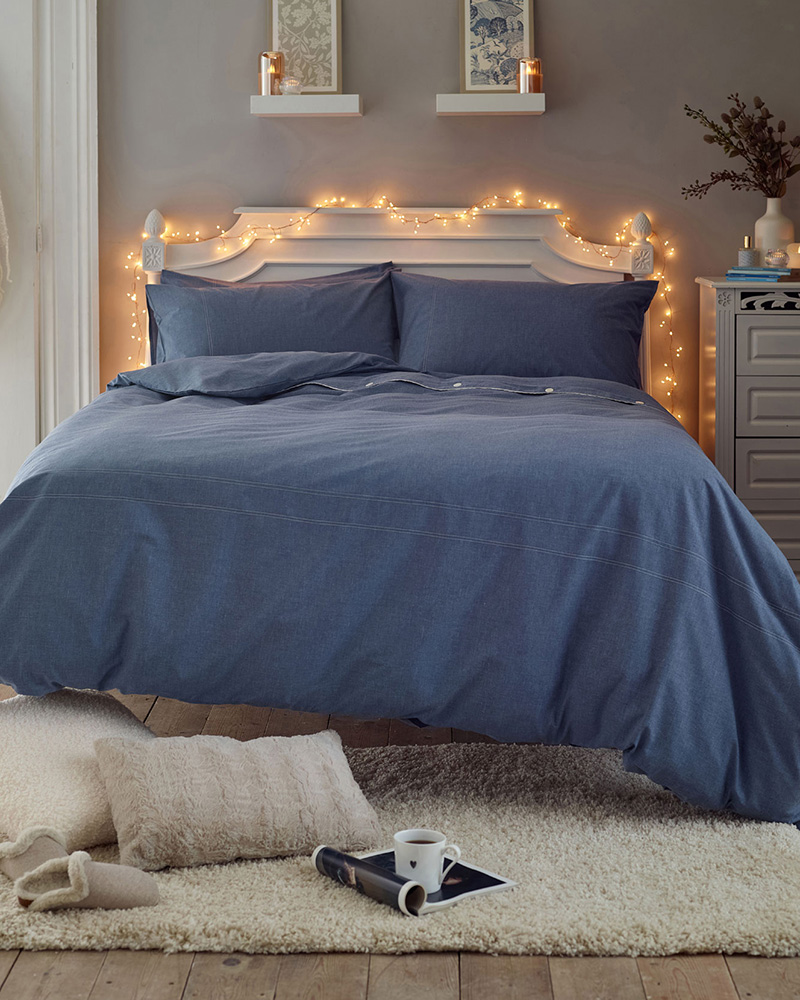 A bedroom with a bed featuring blue floral bedding, a side table with flowers, a hanging light, and a window with plants.