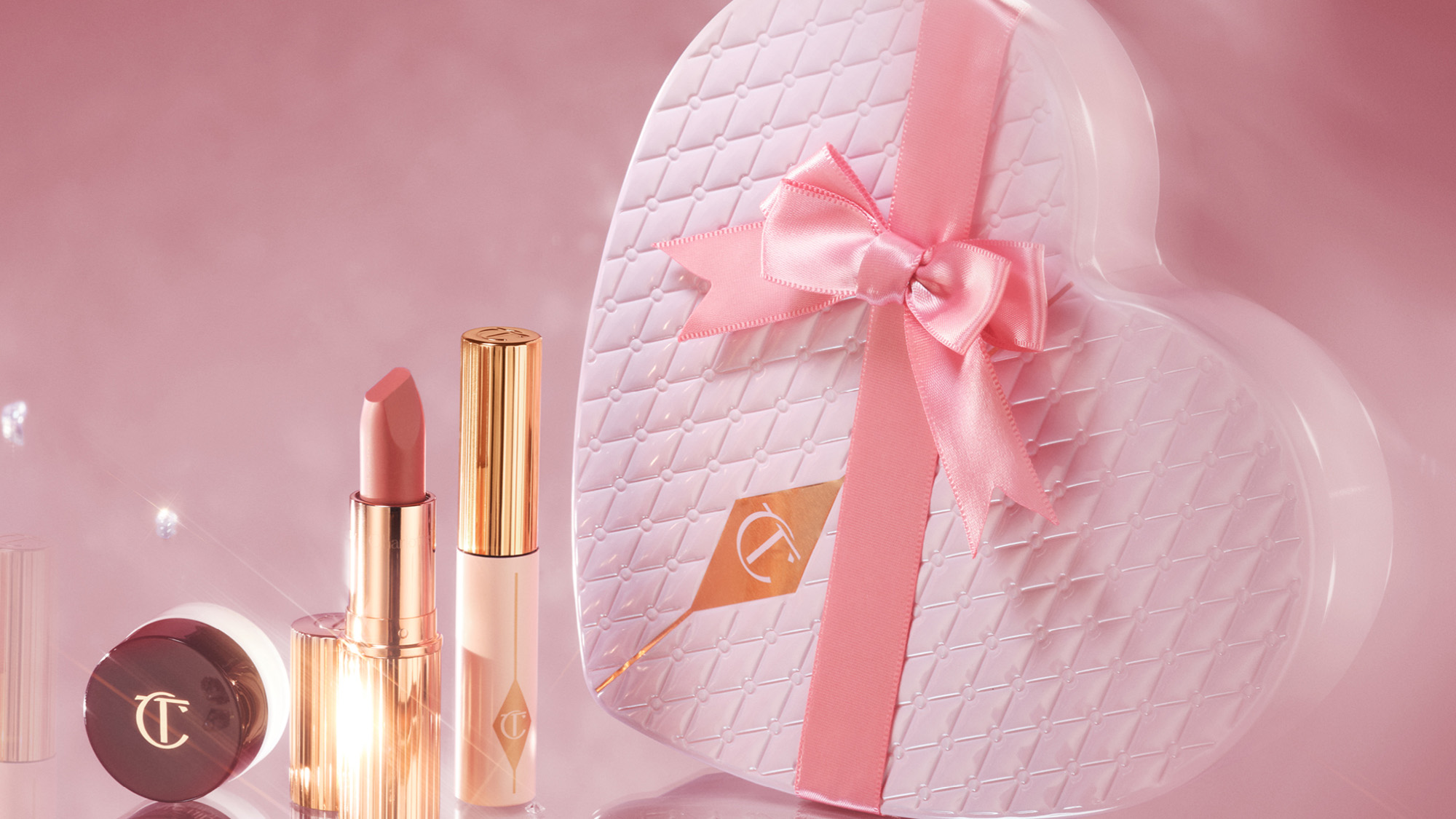 Makeup products with gold accents and a heart-shaped pink gift box with a bow on a soft pink background.