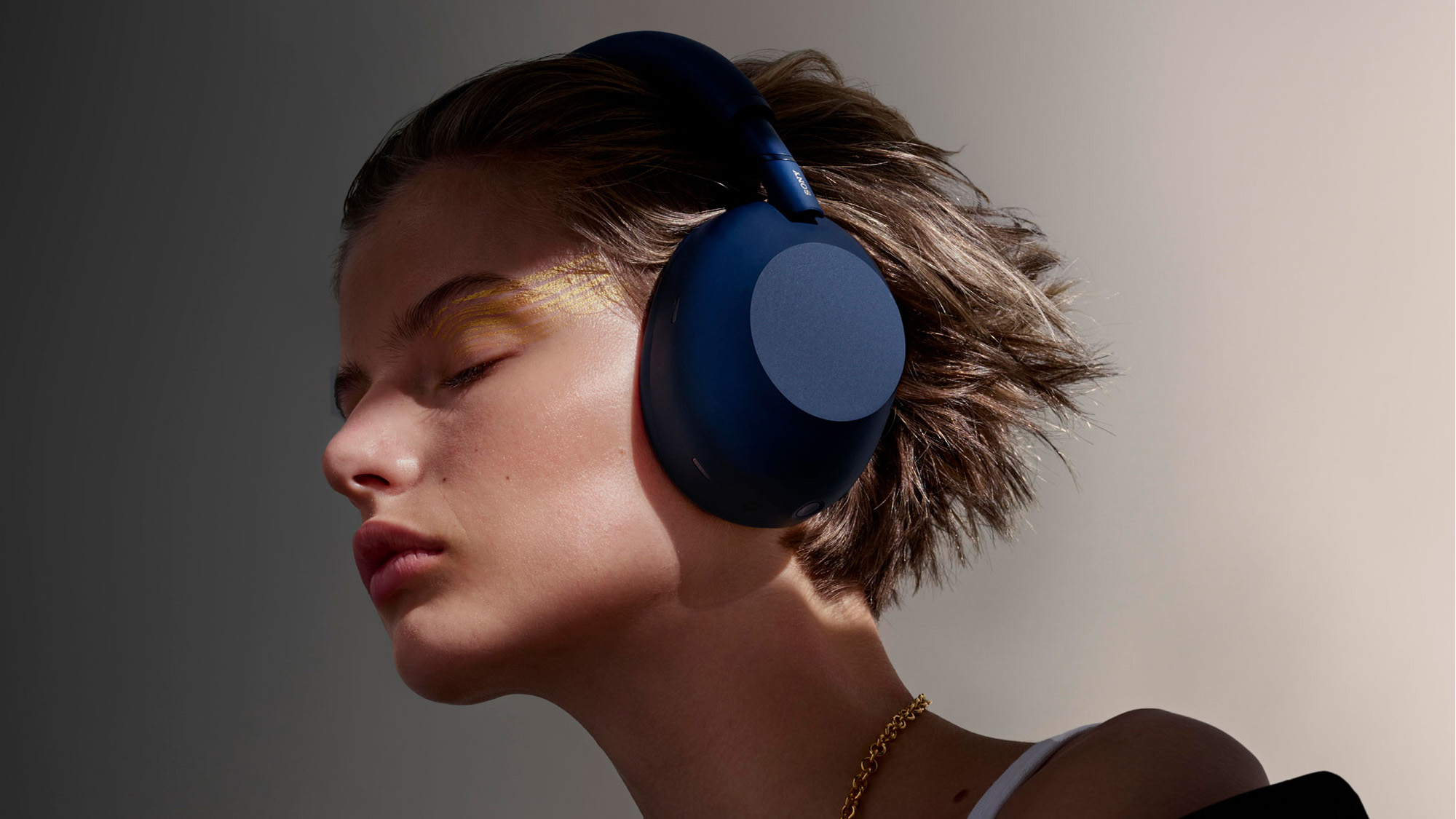 Person wearing large blue headphones, eyes closed, and dressed in a jacket and necklace, bathed in soft lighting.