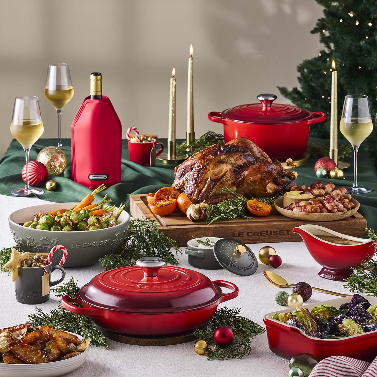 Festive holiday table with roast turkey, side dishes, candles, wine glasses, ornaments, and cookware.