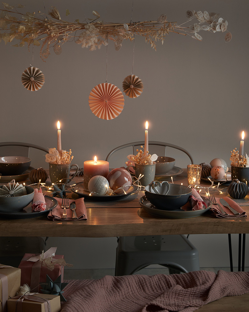 A dimly lit dining table is set with dishes, candles, and greenery. A person in a light blouse is adjusting a candle.