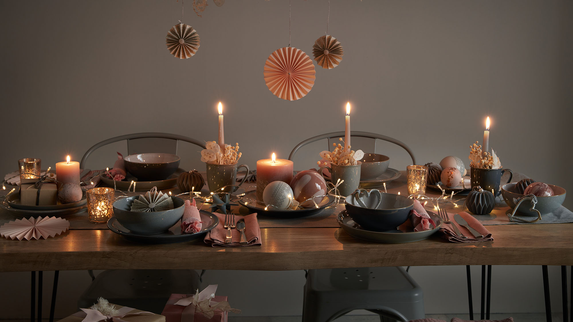 A dimly lit dining table is set with dishes, candles, and greenery. A person in a light blouse is adjusting a candle.