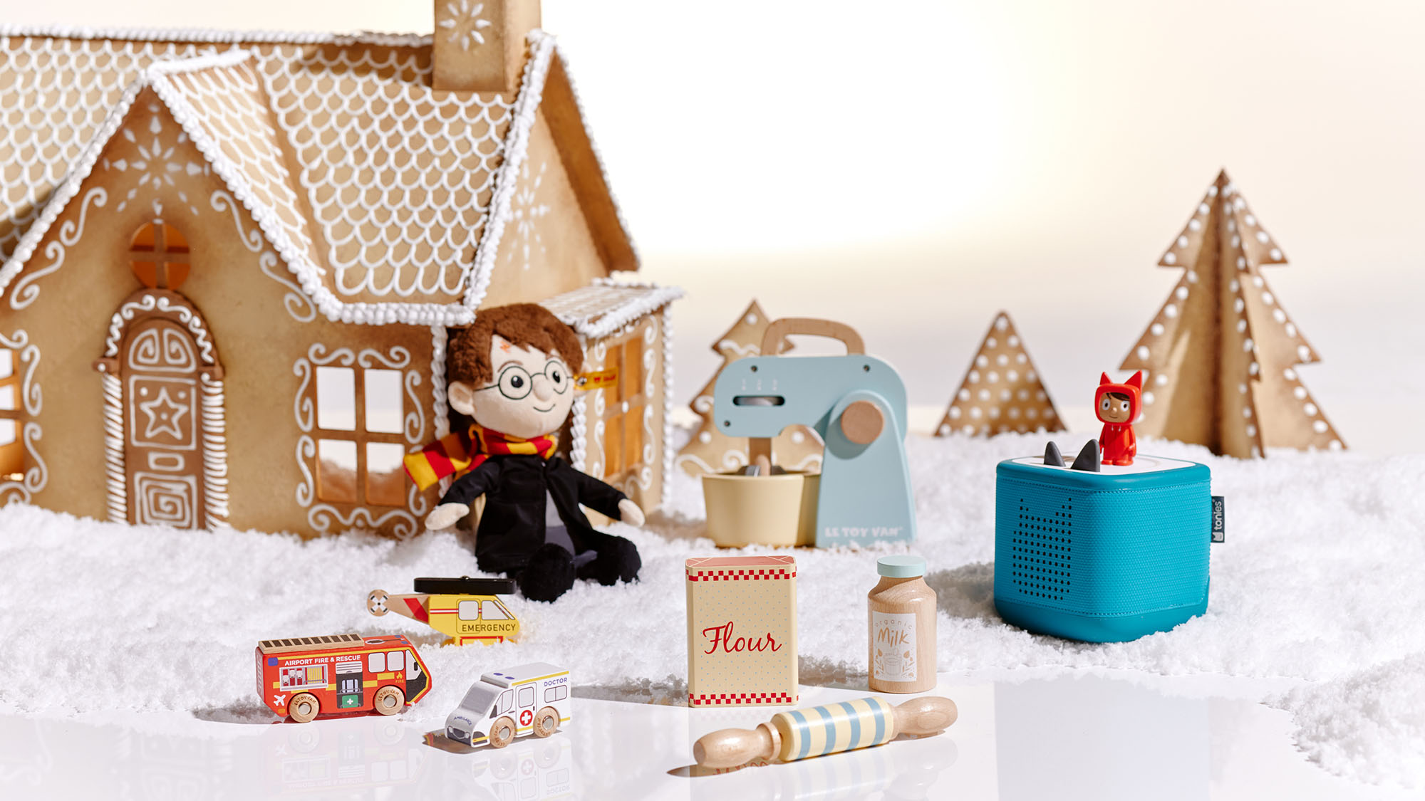 A gingerbread house scene with toy figures, a doll, and miniature kitchen utensils, set on a snow-like surface.