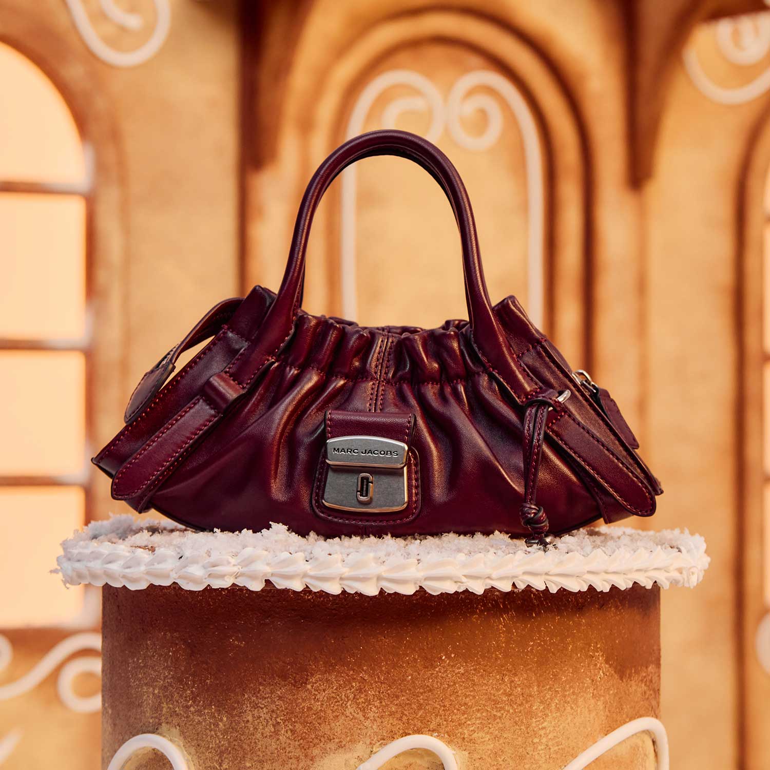 A stylish burgundy handbag sits on a decorative cake-like stand with an ornate gingerbread house background.