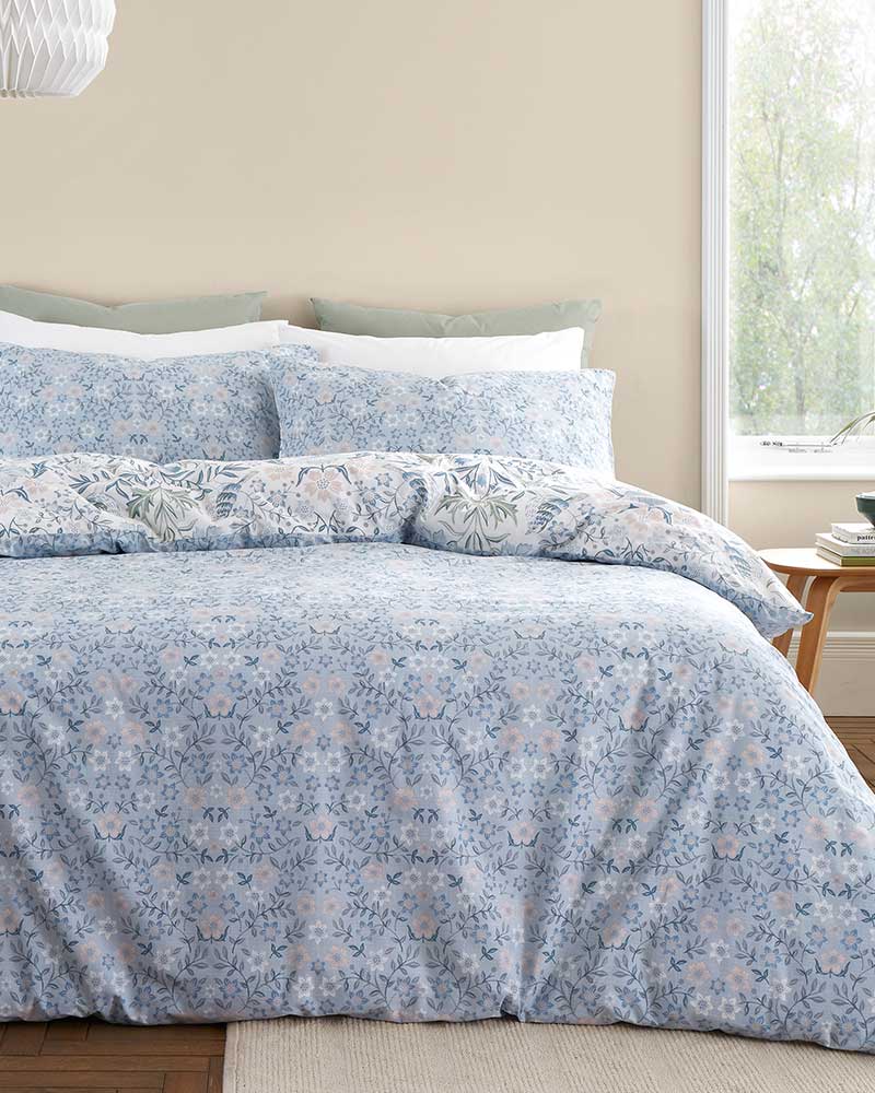 A bed with floral-patterned blue bedding, pillows, and a bedside table near a window.