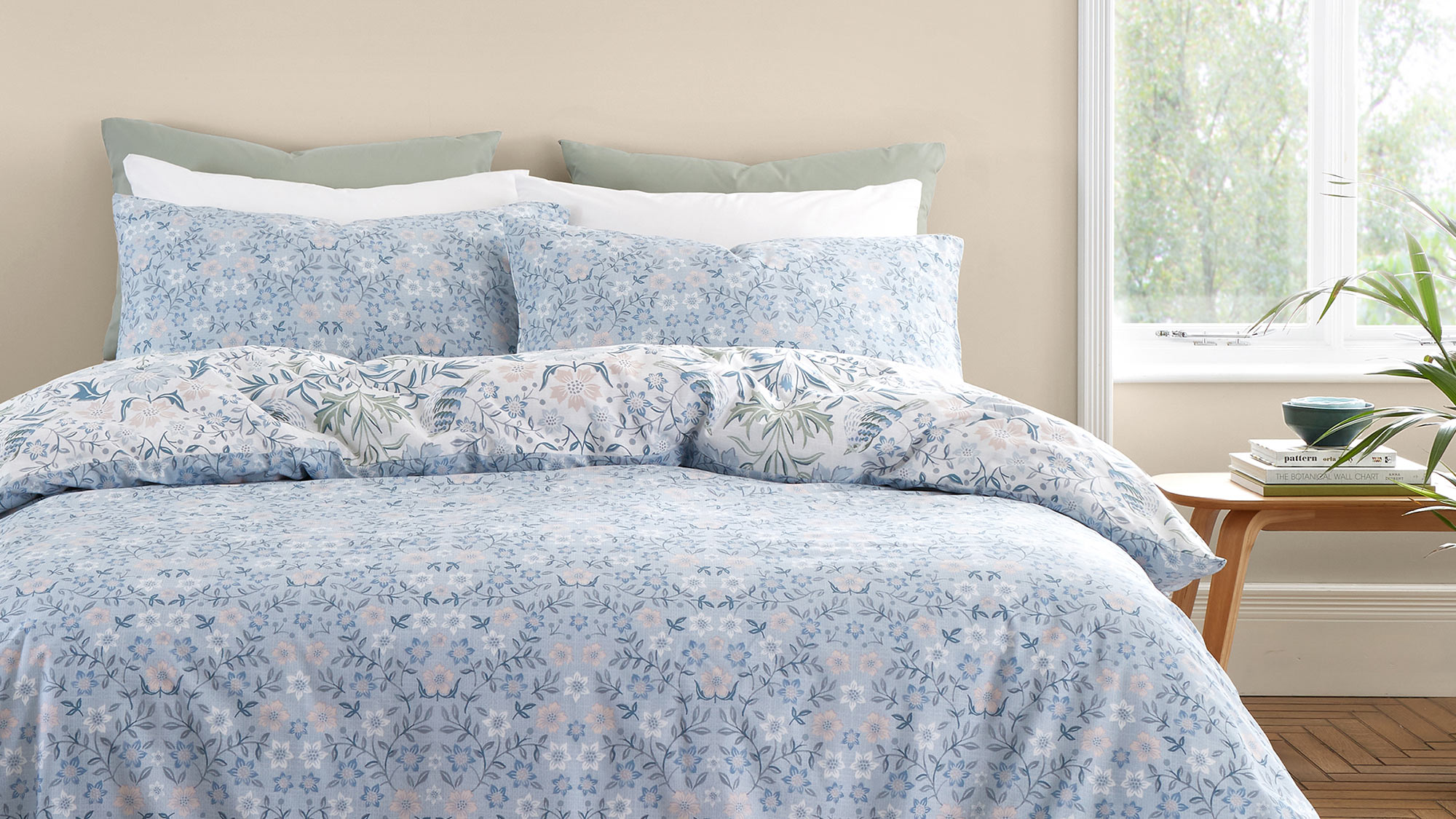 A bed with floral-patterned blue bedding, pillows, and a bedside table near a window.