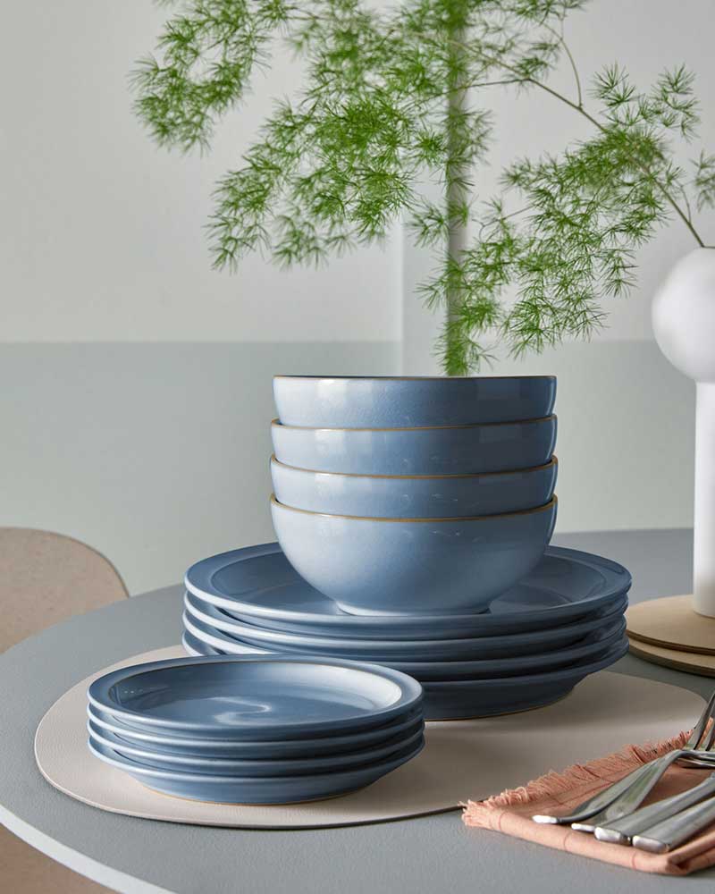 Stacked blue dishes and bowls on a table with cutlery, a napkin, and a vase with green foliage.
