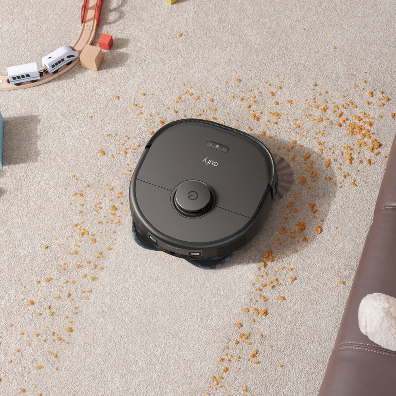 A robot vacuum cleaning crumbs on a carpet near a toy train and large foam blocks.