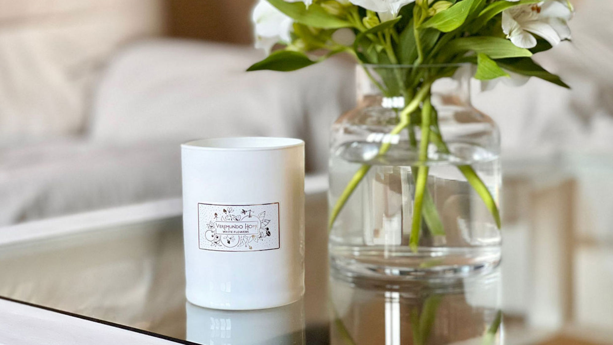 A white candle and a clear vase of white flowers on a glass table.
