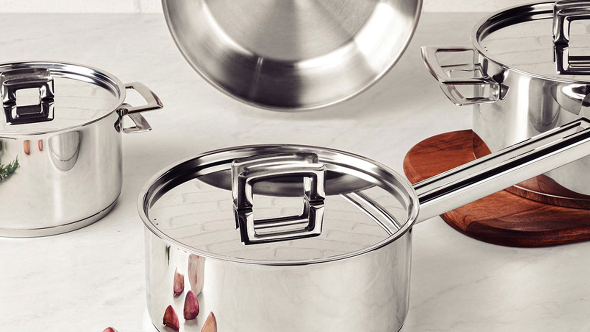 Stainless steel cookware set on a kitchen counter, with garlic cloves nearby.