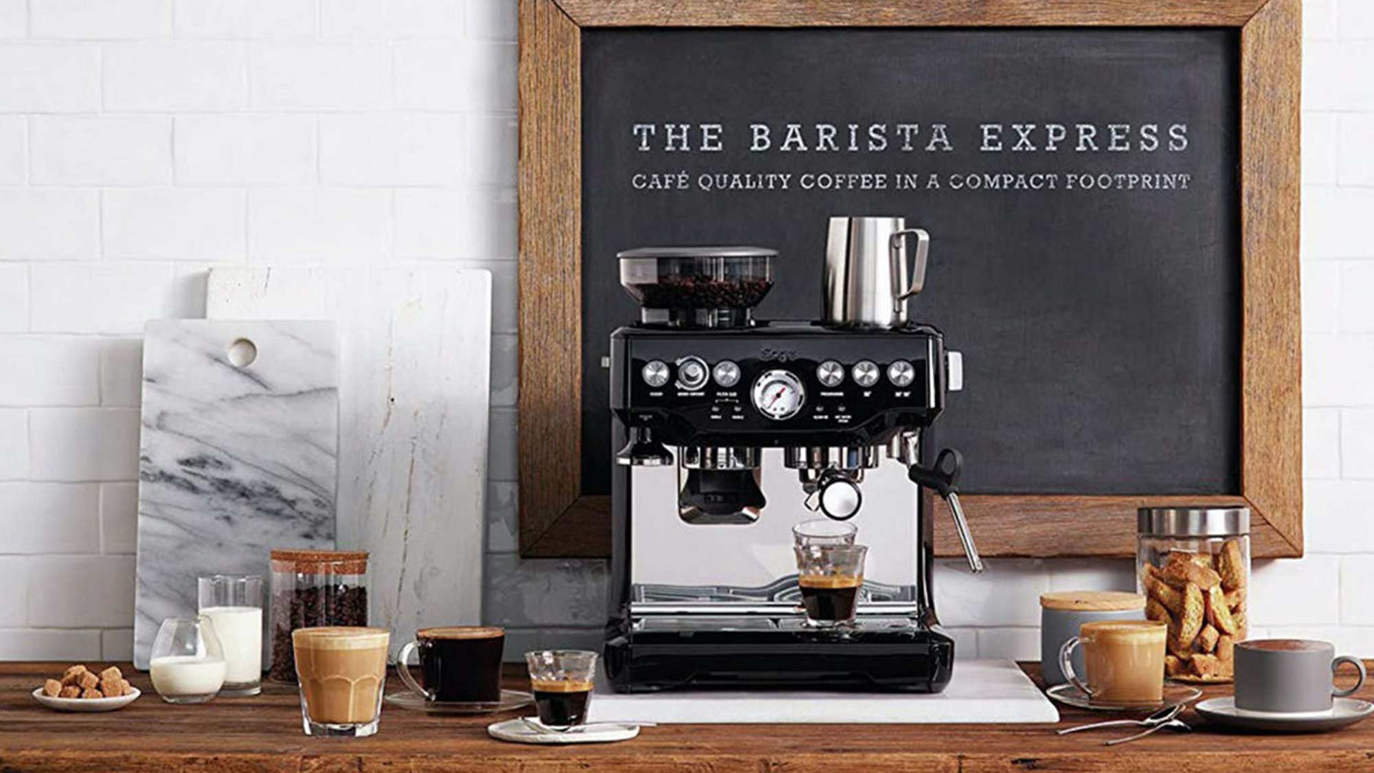 A coffee machine with assorted cups and biscuits is set against a white tiled wall with a framed sign reading 'The Barista Express.'