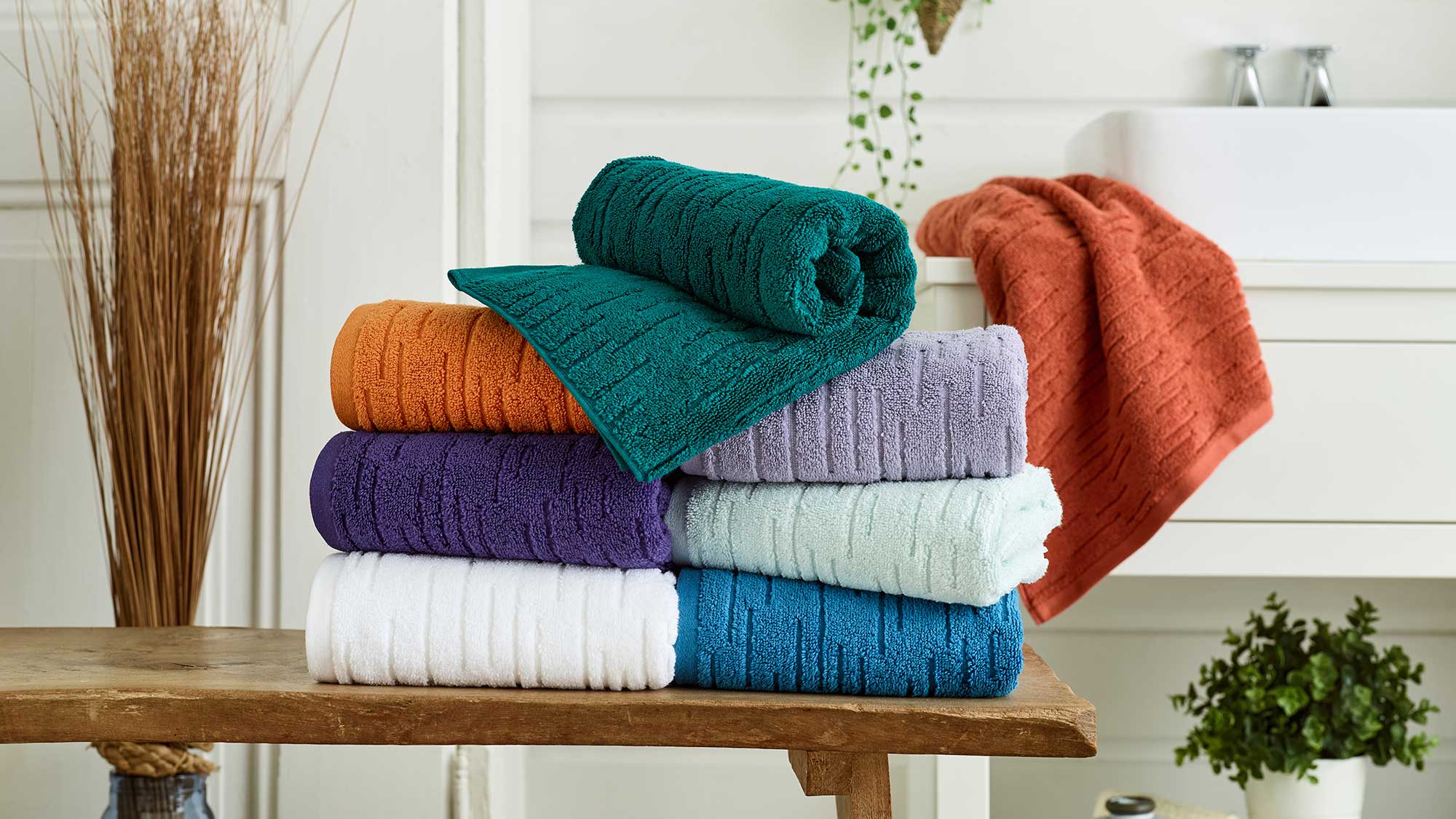Stack of colorful towels on a wooden bench in a bathroom with plants and decor.