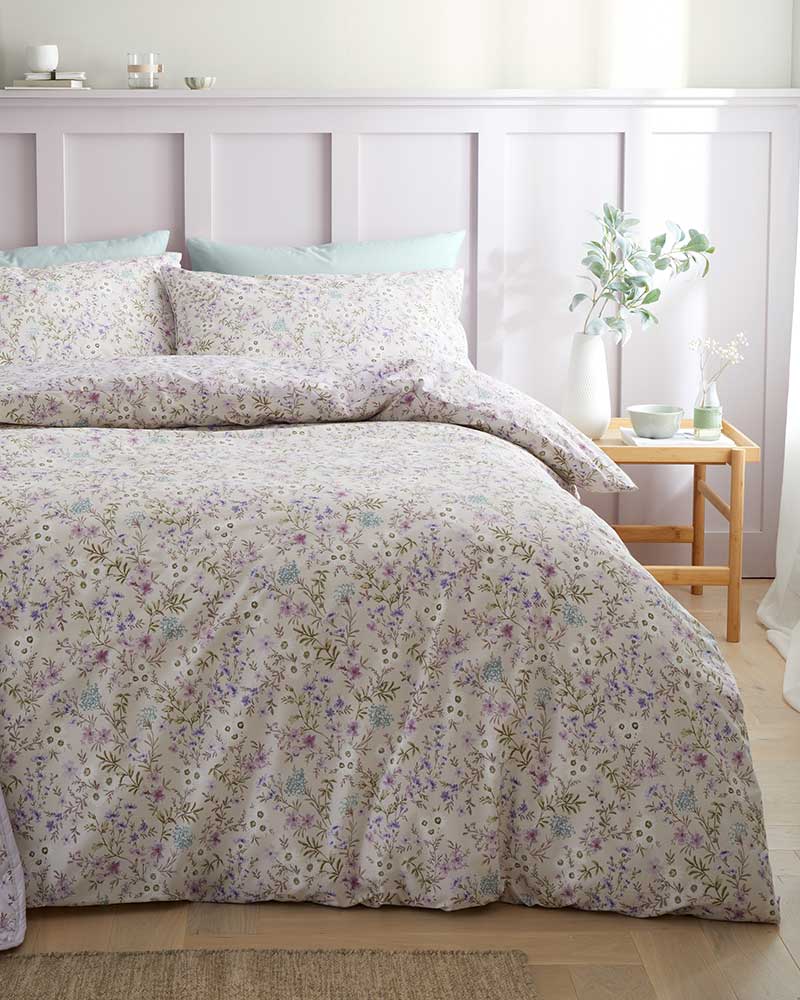 Floral-patterned bedding set with pillows on a bed, wooden side table with decor, and part of a light pink paneled wall.