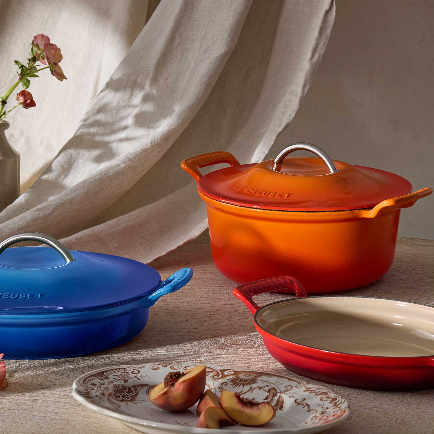 Colorful cookware with lids on a table, flowers, and bottles in the background.