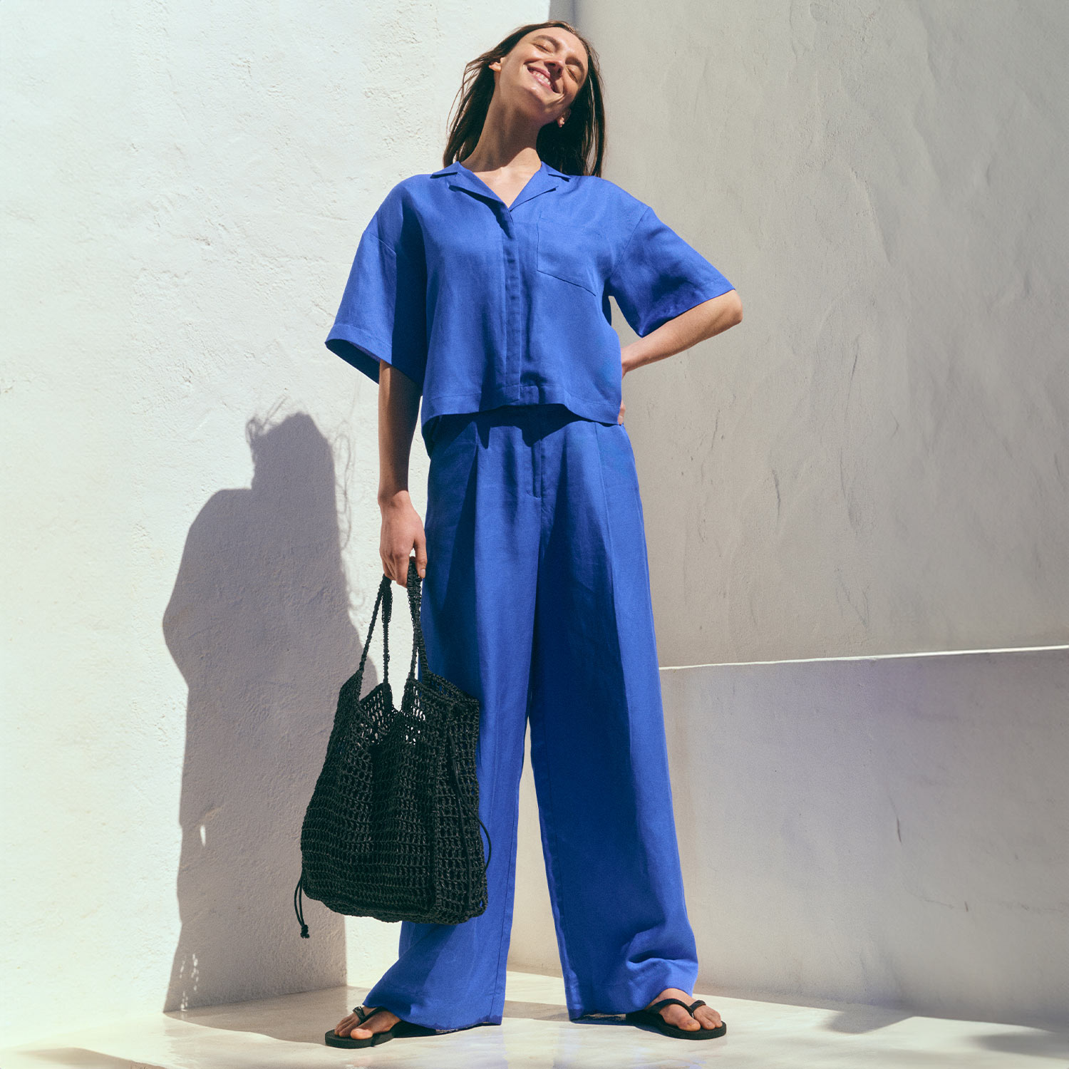 A person in a bright blue outfit smiles under the sun, holding a black woven bag and standing against a white background.